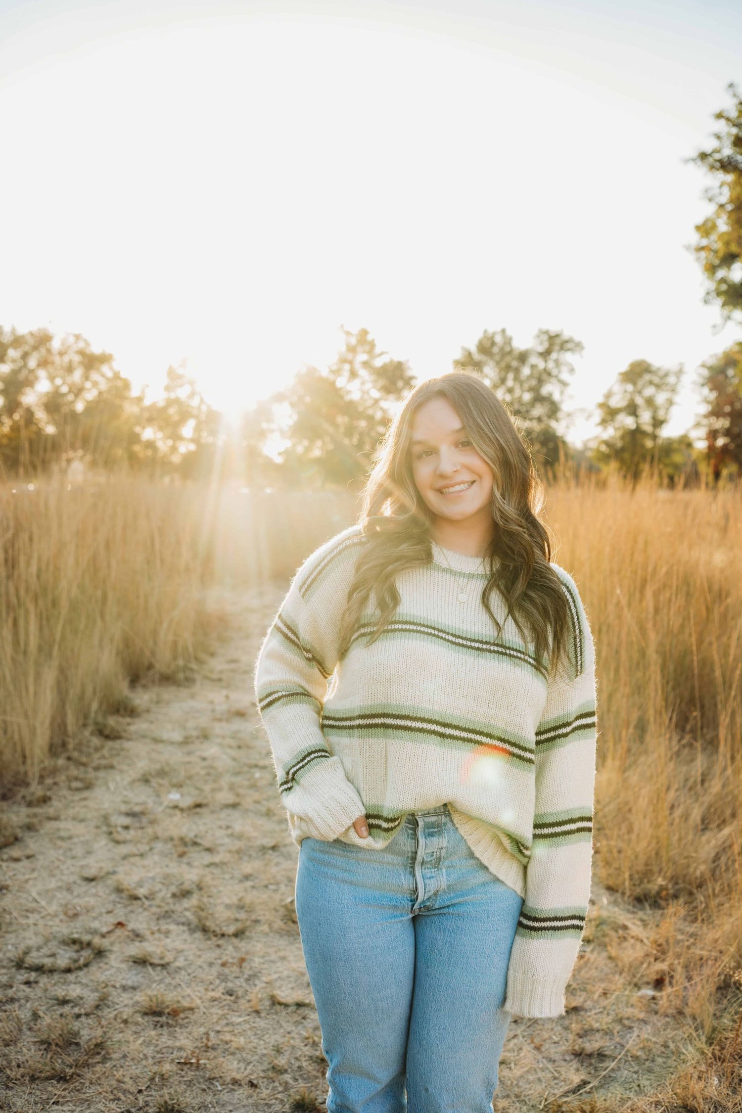 A senior portrait taken during golden hour of a girl wearing a sweater and jeans with one hand in her pocket and a soft smile on her face.