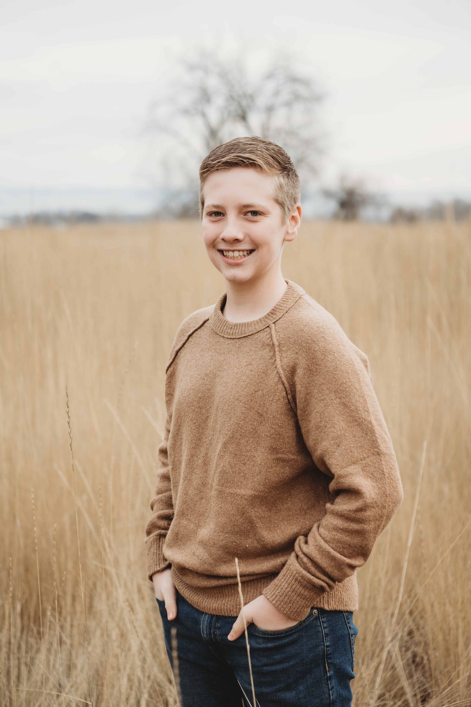 A teenage boy wearing neutral tones smiling with his hands in his pockets.