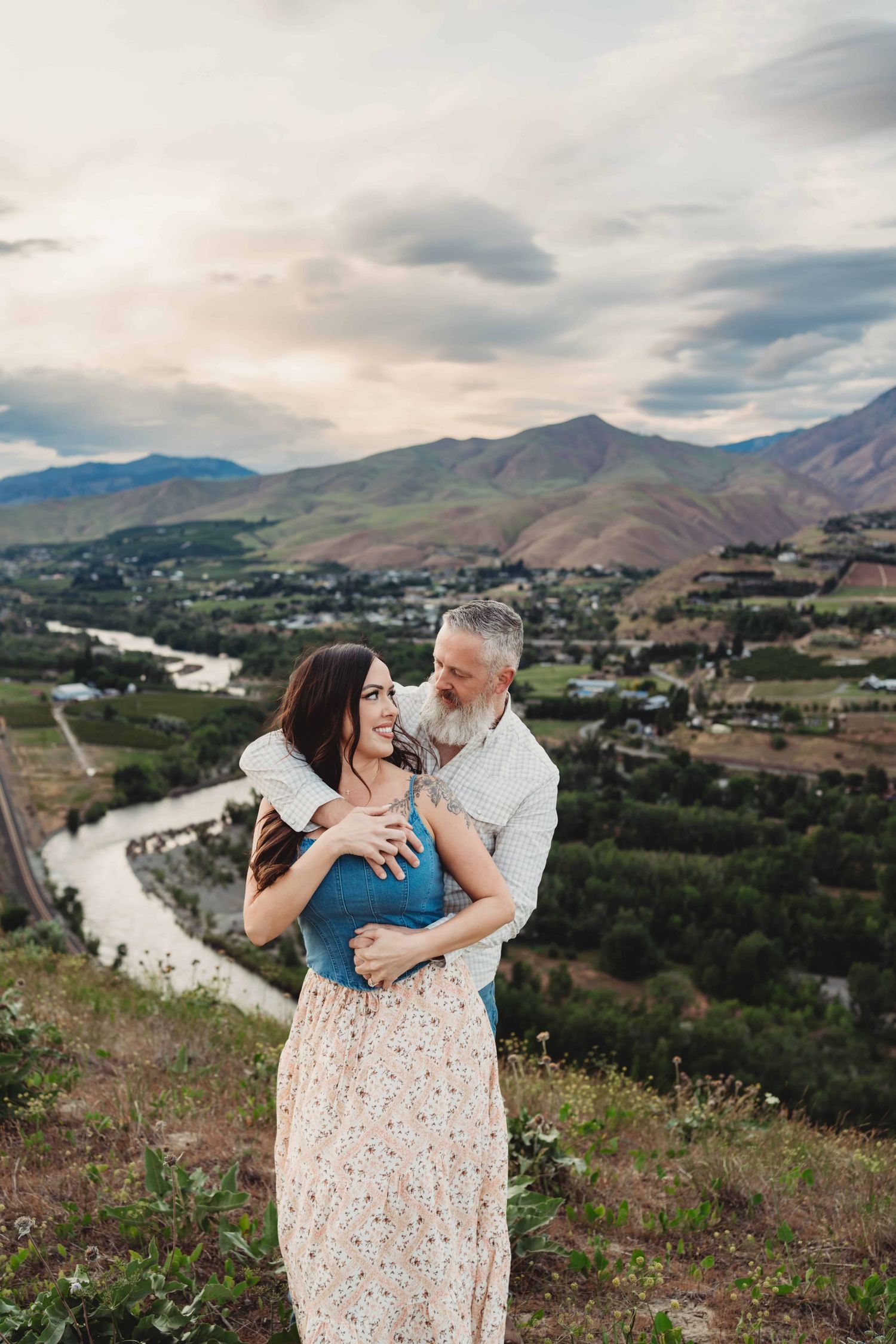 A couple standing above a river with mountains and a cloudy sunset in the background.