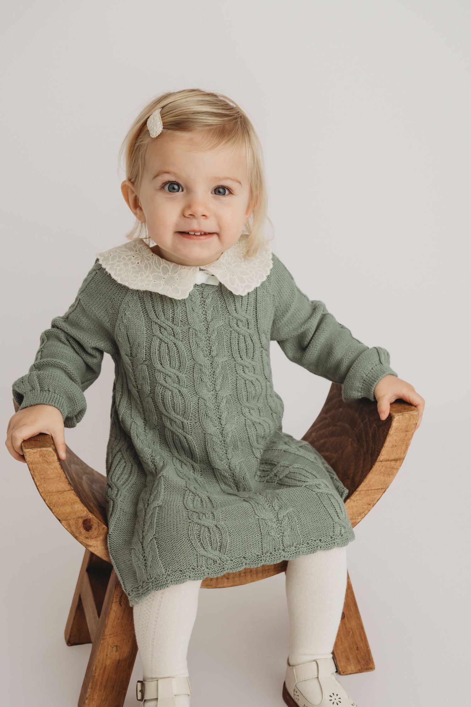 A one year old girl sitting on a wooden stool in a vintage green dress.