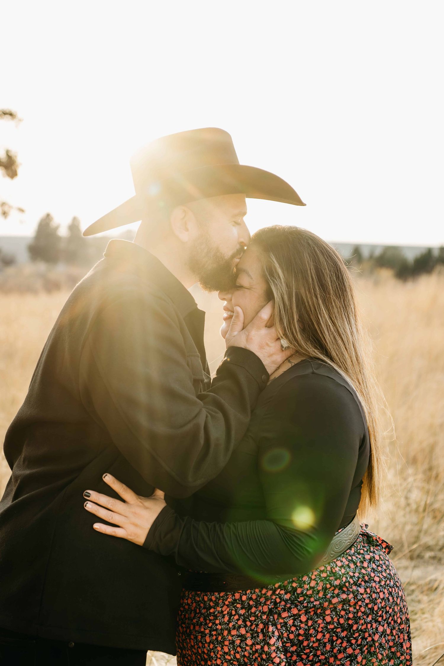 A golden light filled portrait of a man wearing a cowboy hat kissing his girlfriend's forehead.