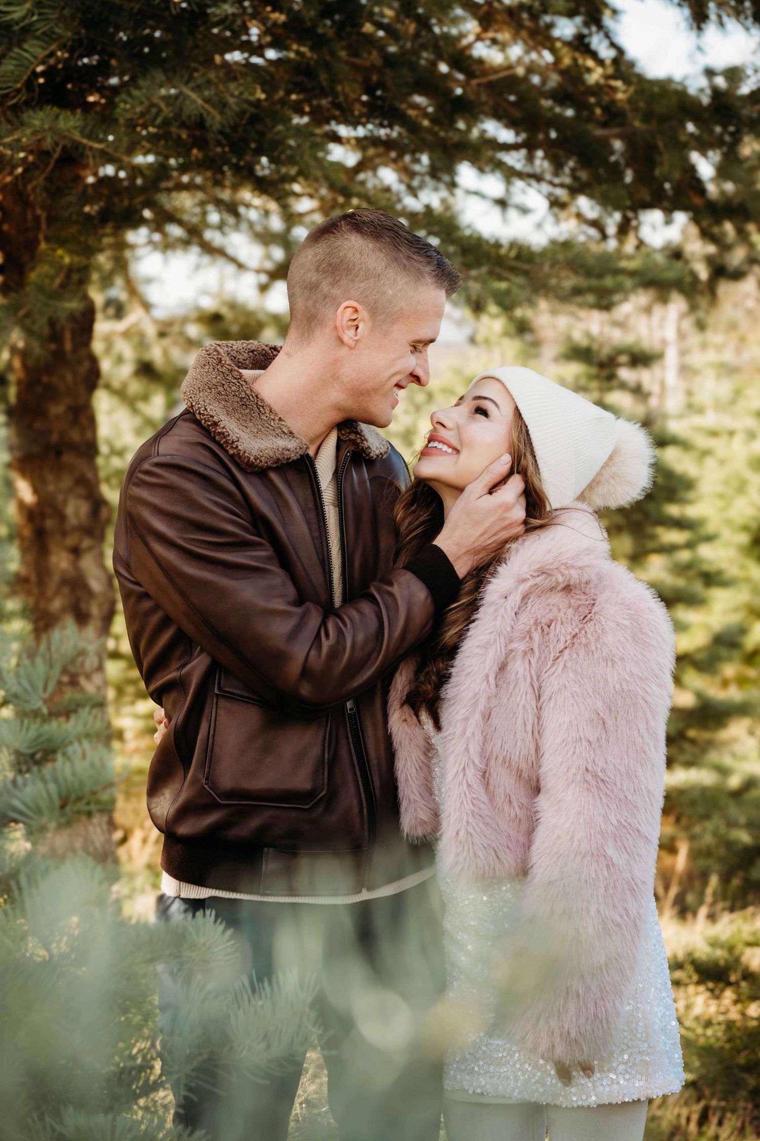 A couple surrounded by evergreen trees looking at each other dressed in warm winter clothing.