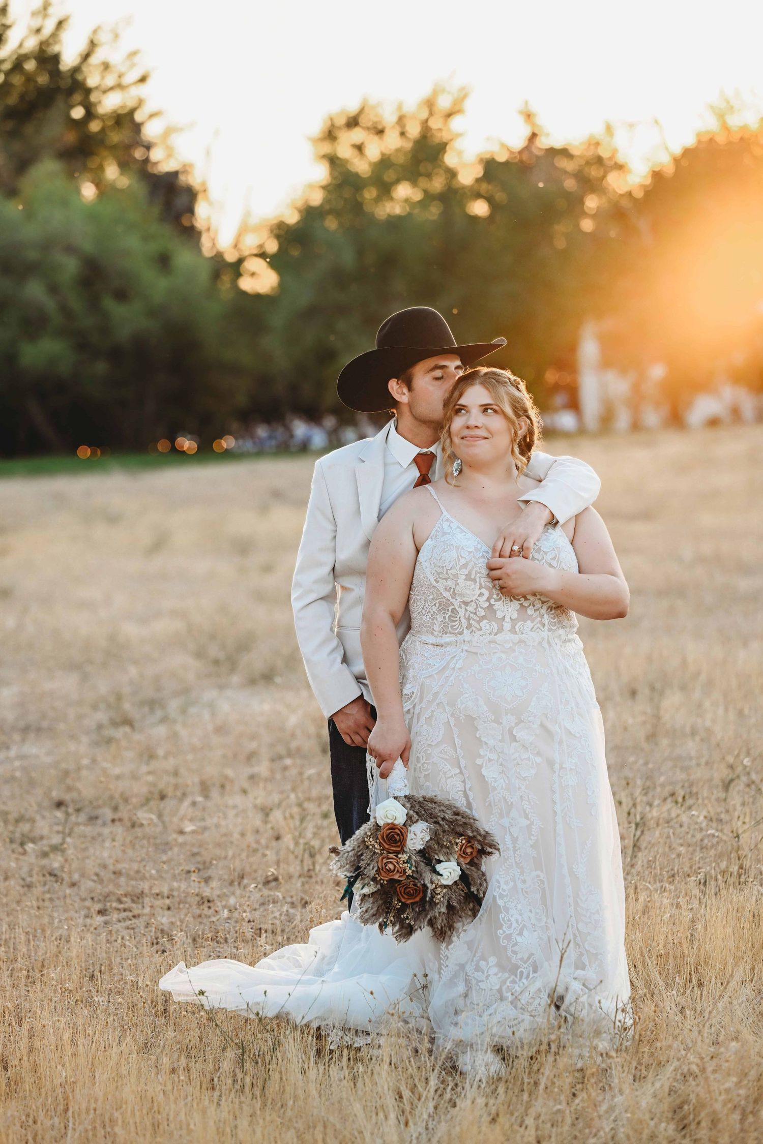 A couple standing in a field during golden hour while a man wearing a cowboy hat kisses his bride's head.