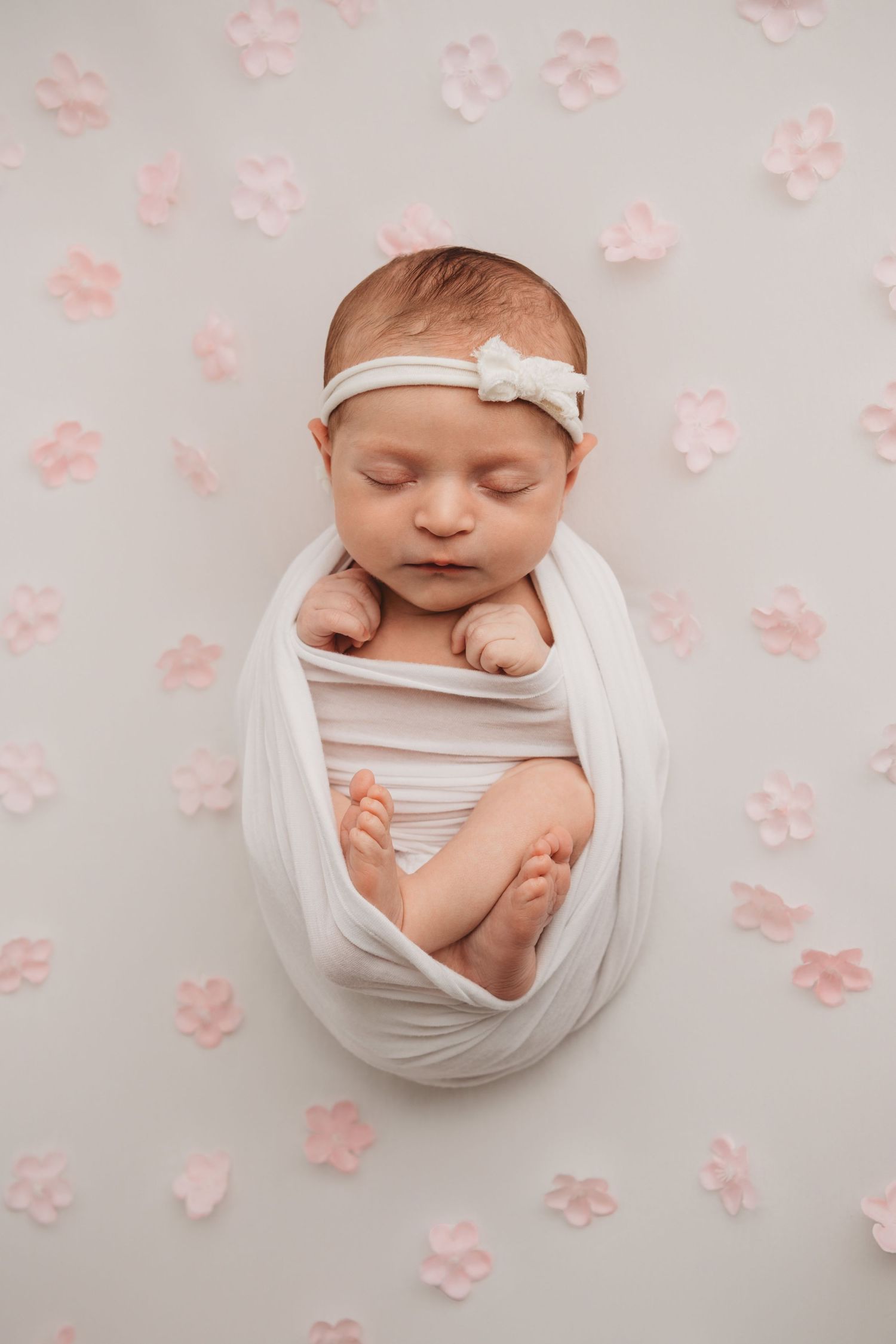 A portrait of a newborn girl wrapped in white with pink flowers surrounding her.