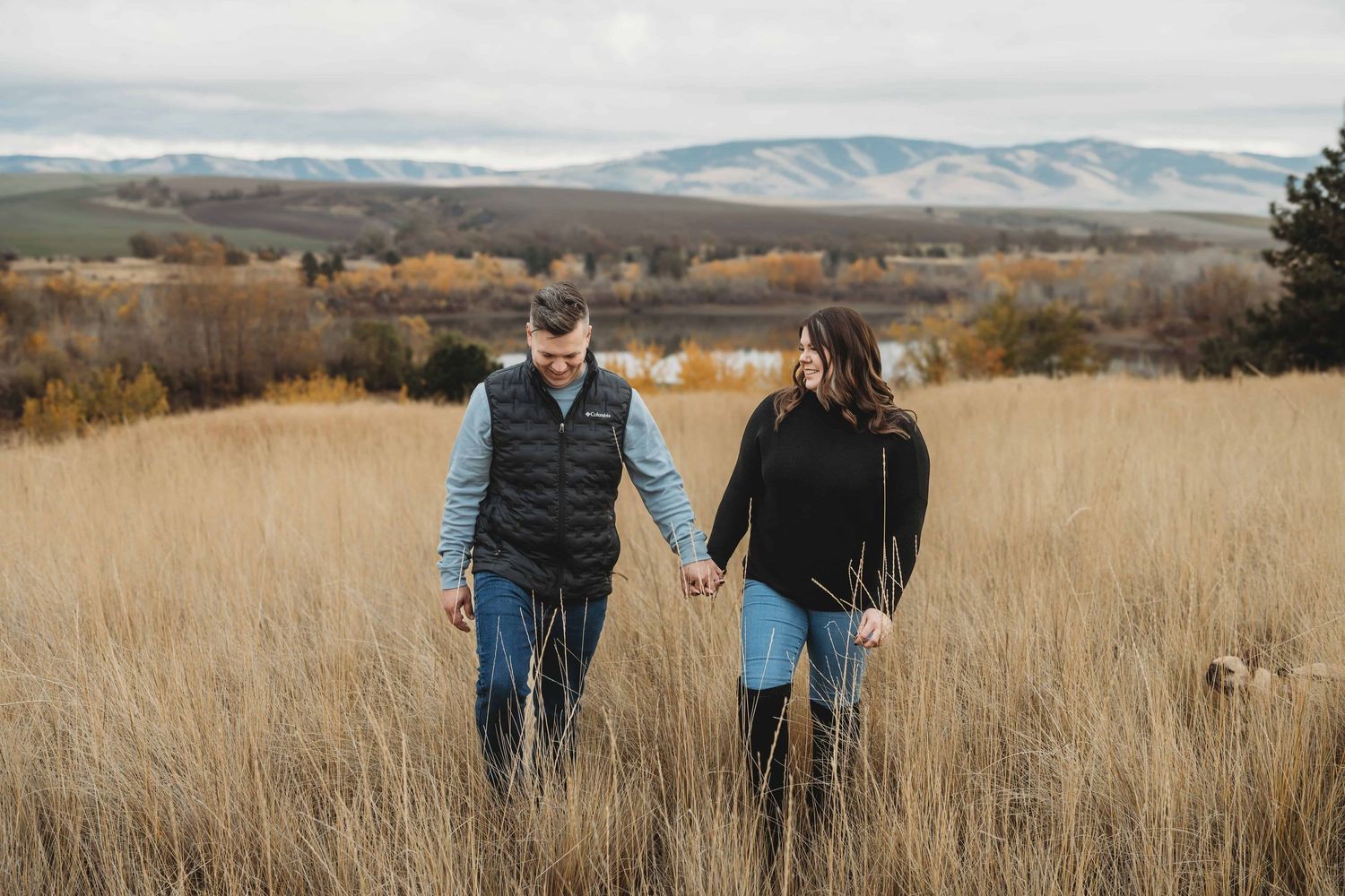A portrait of a couple walking in a field through tall grasses with a lake and mountains in the background.