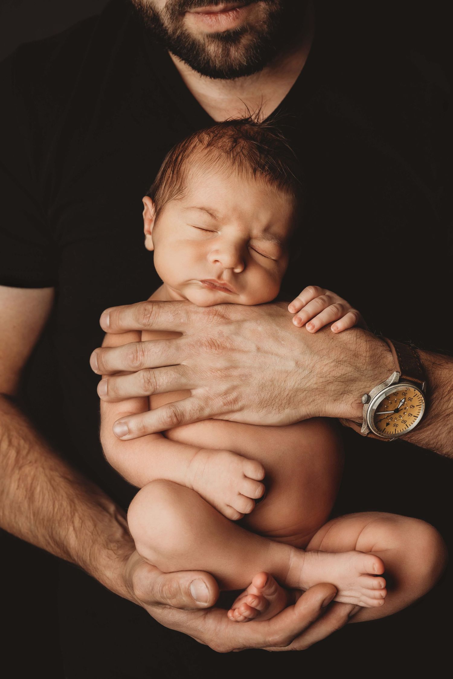 Dark and moody studio portrait of a sleeping newborn baby in his father's arms.