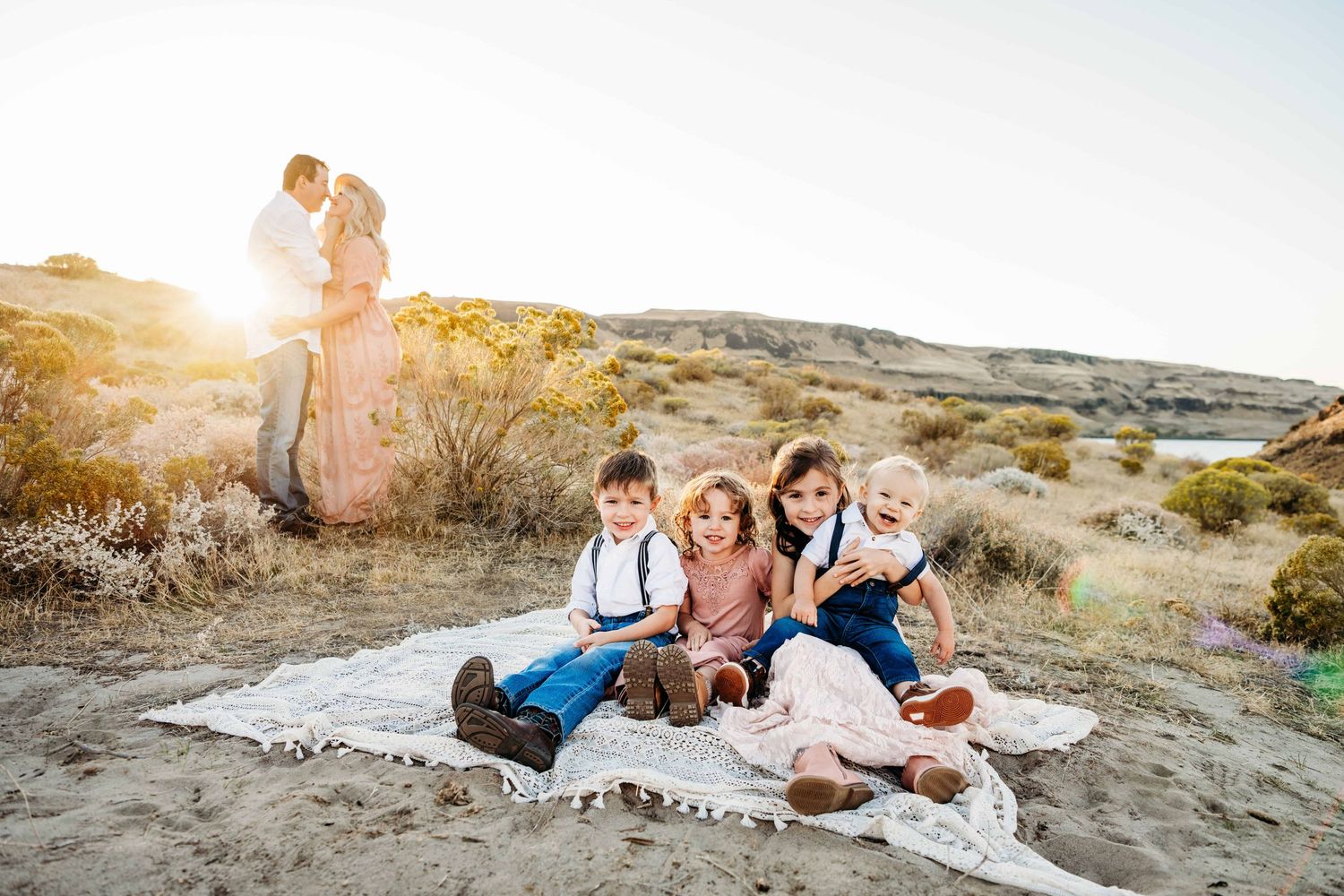 Children sit on a blanket in the foreground while their parents kiss in the background.
