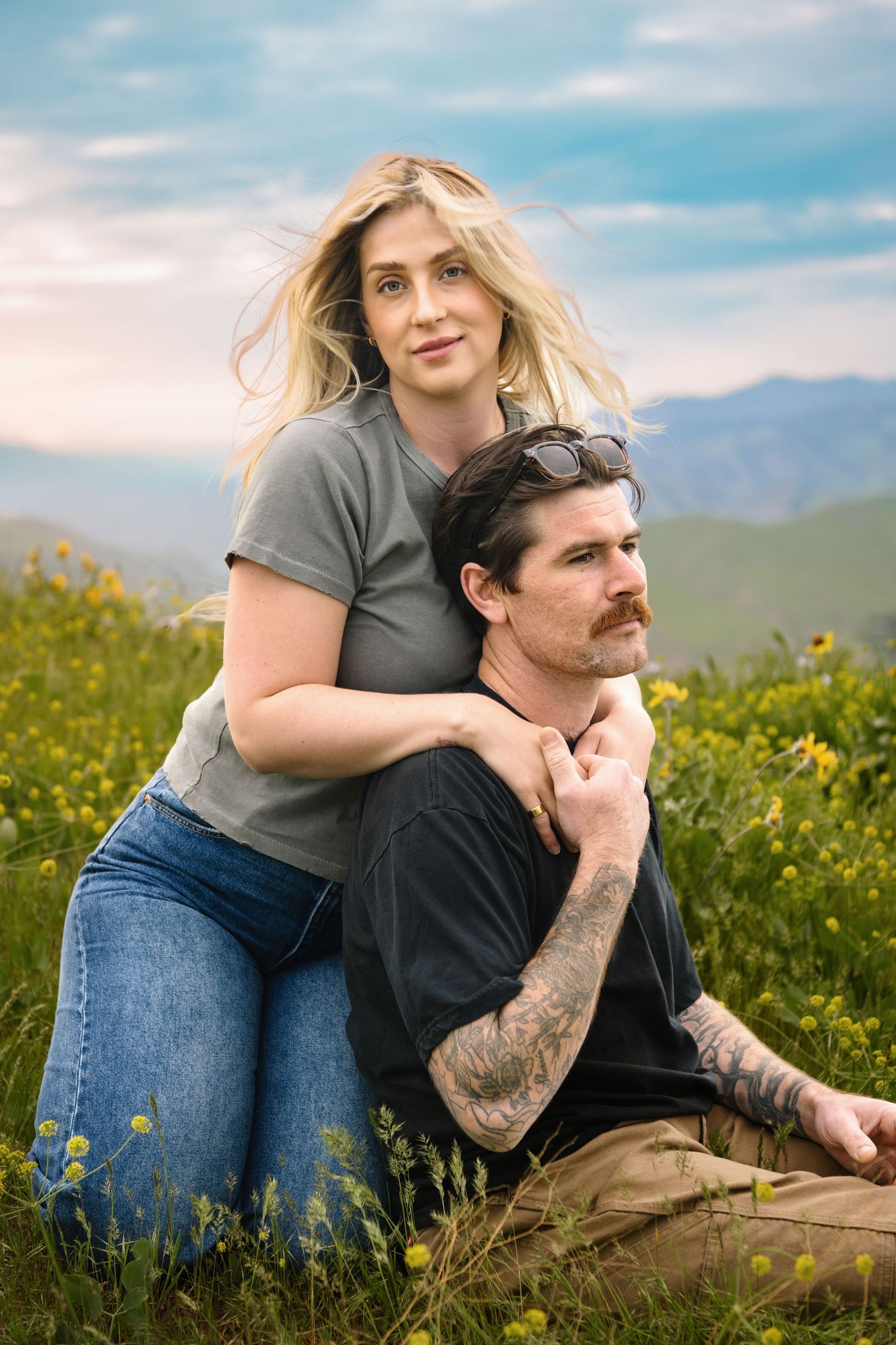 A couple sitting in a wildflower field in Wenatchee while the woman looks at the camera and the man looks off into the distance.