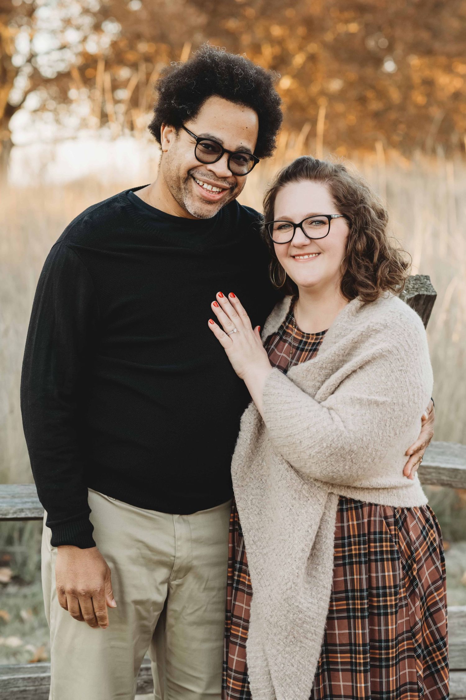 A couple standing in front of an old wooden fence dressed in fall colors with fall colored trees in the background.