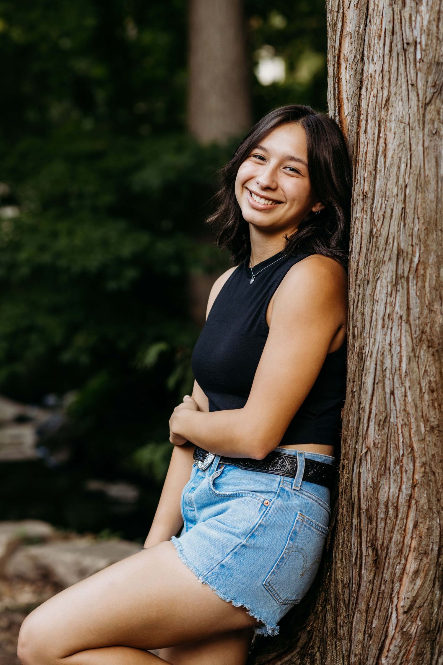 A senior leaning against a tree with her arm across her stomach, smiling at the camera.