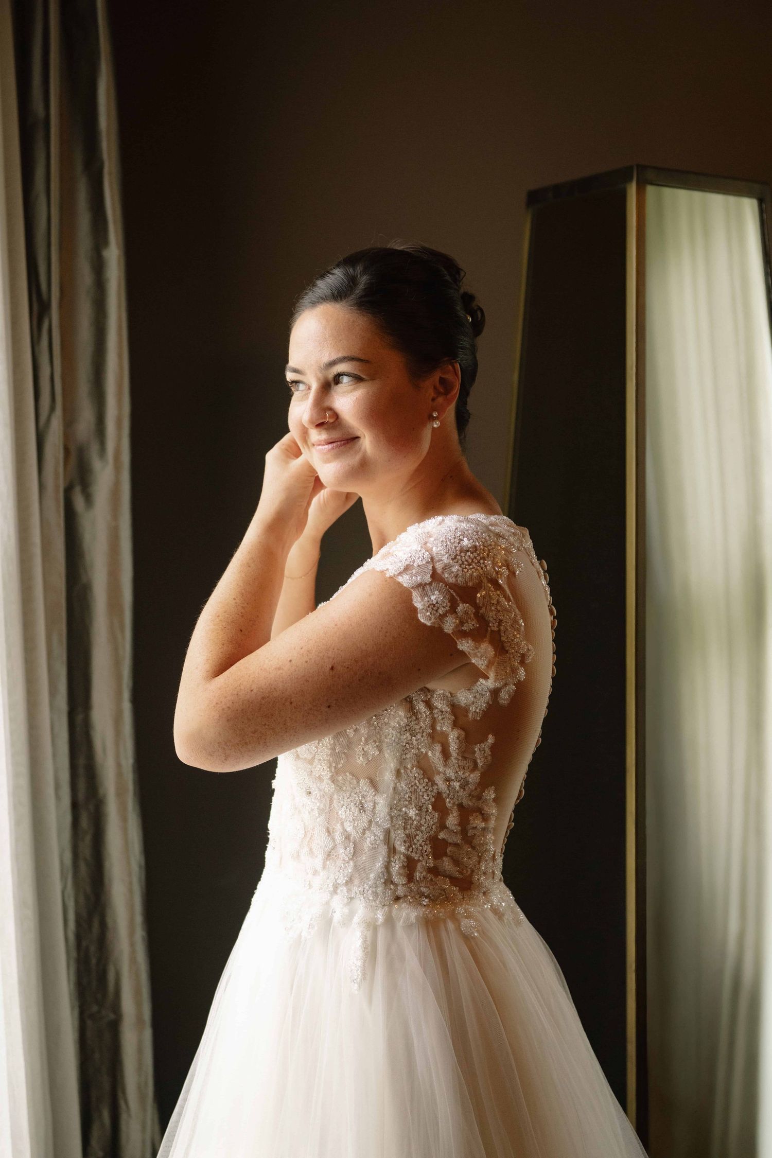 A bride looking through a window with soft lighting while putting in her earrings.