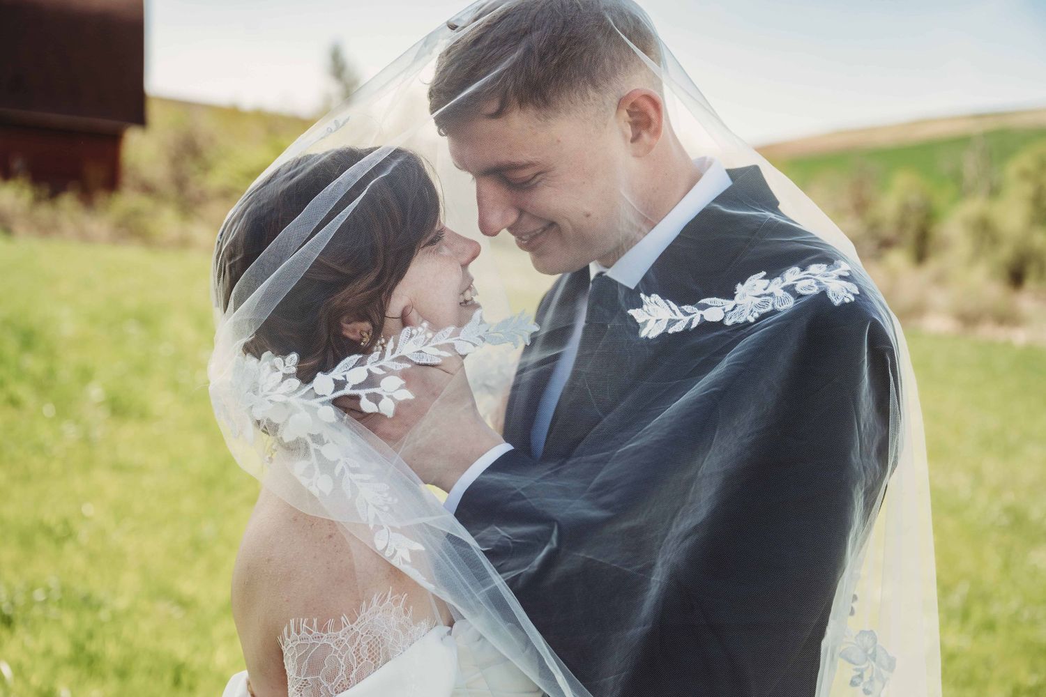 A bride and groom underneath a veil smiling at each other