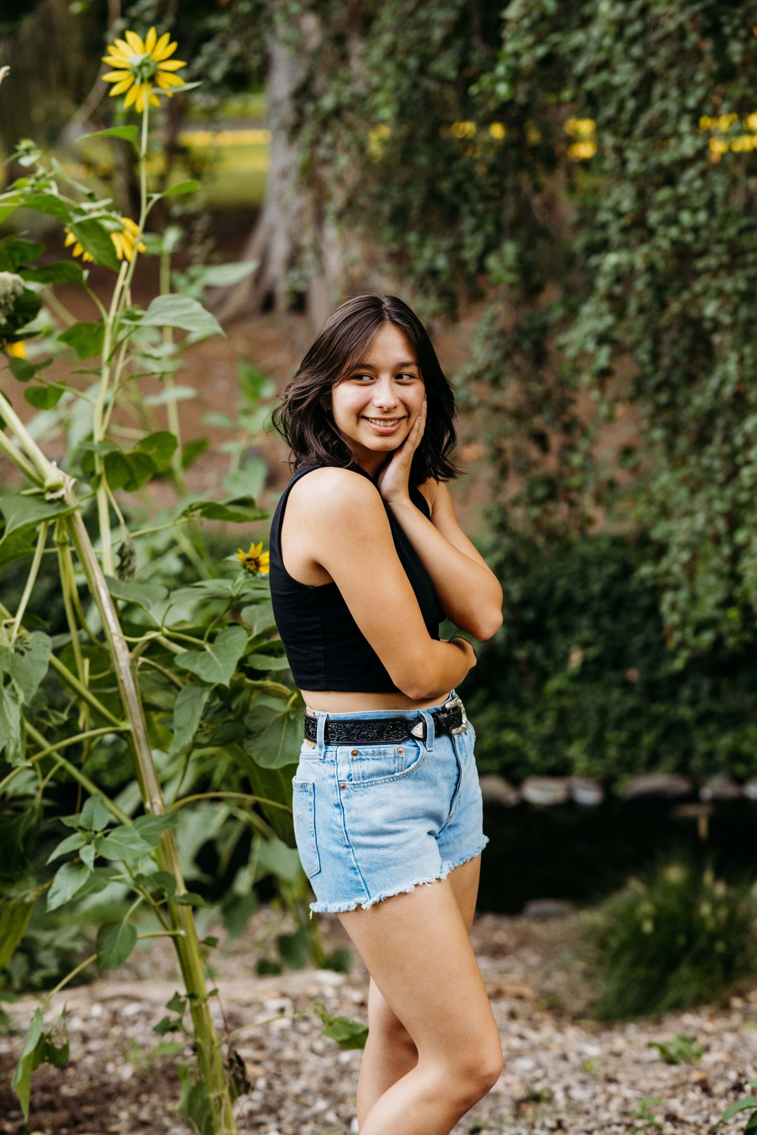 A senior portrait of a girl standing near sunflowers dressed in a black shirt and jean shorts.