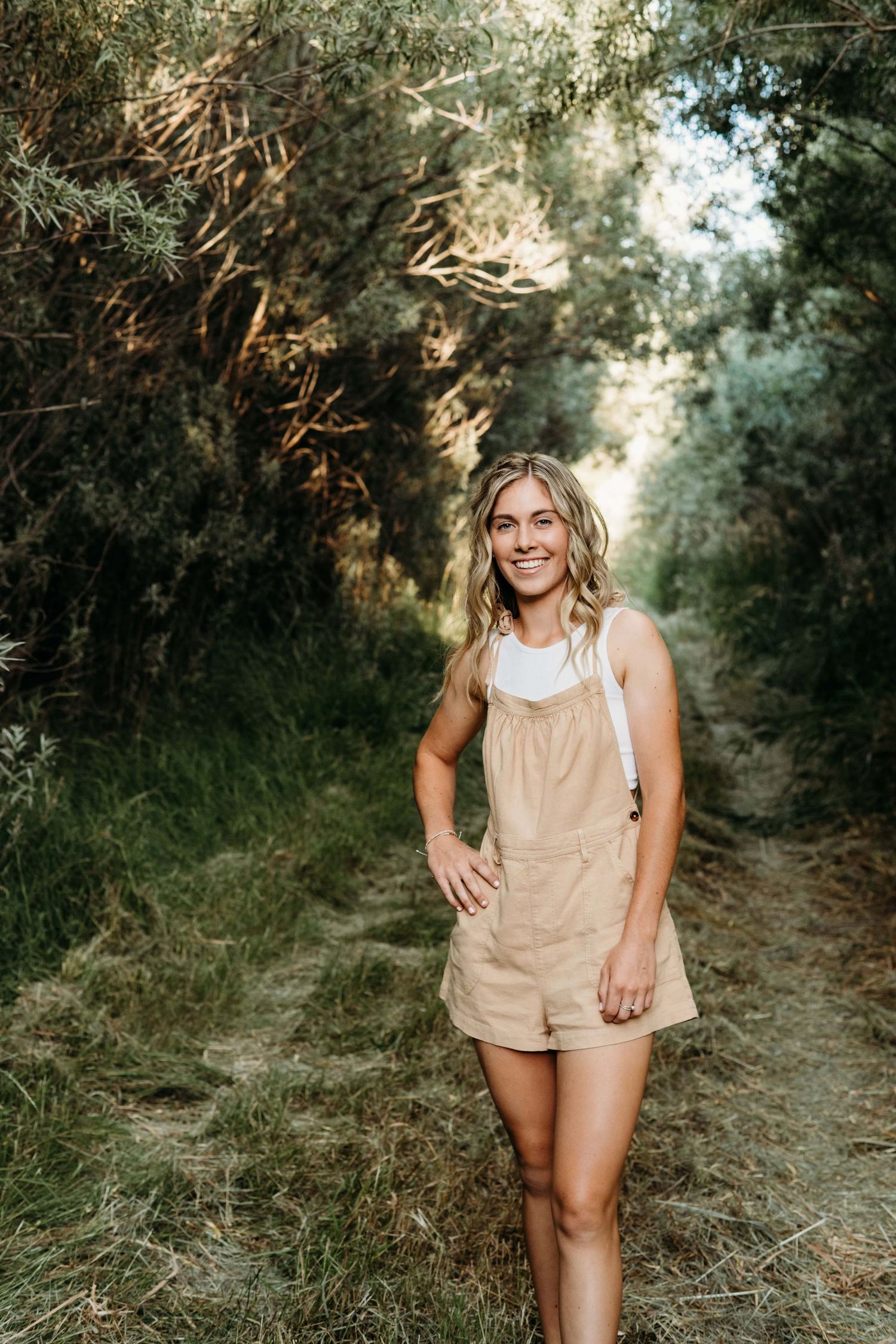A senior standing in a dark tree-covered area with her hand on her hip, smiling for the camera.