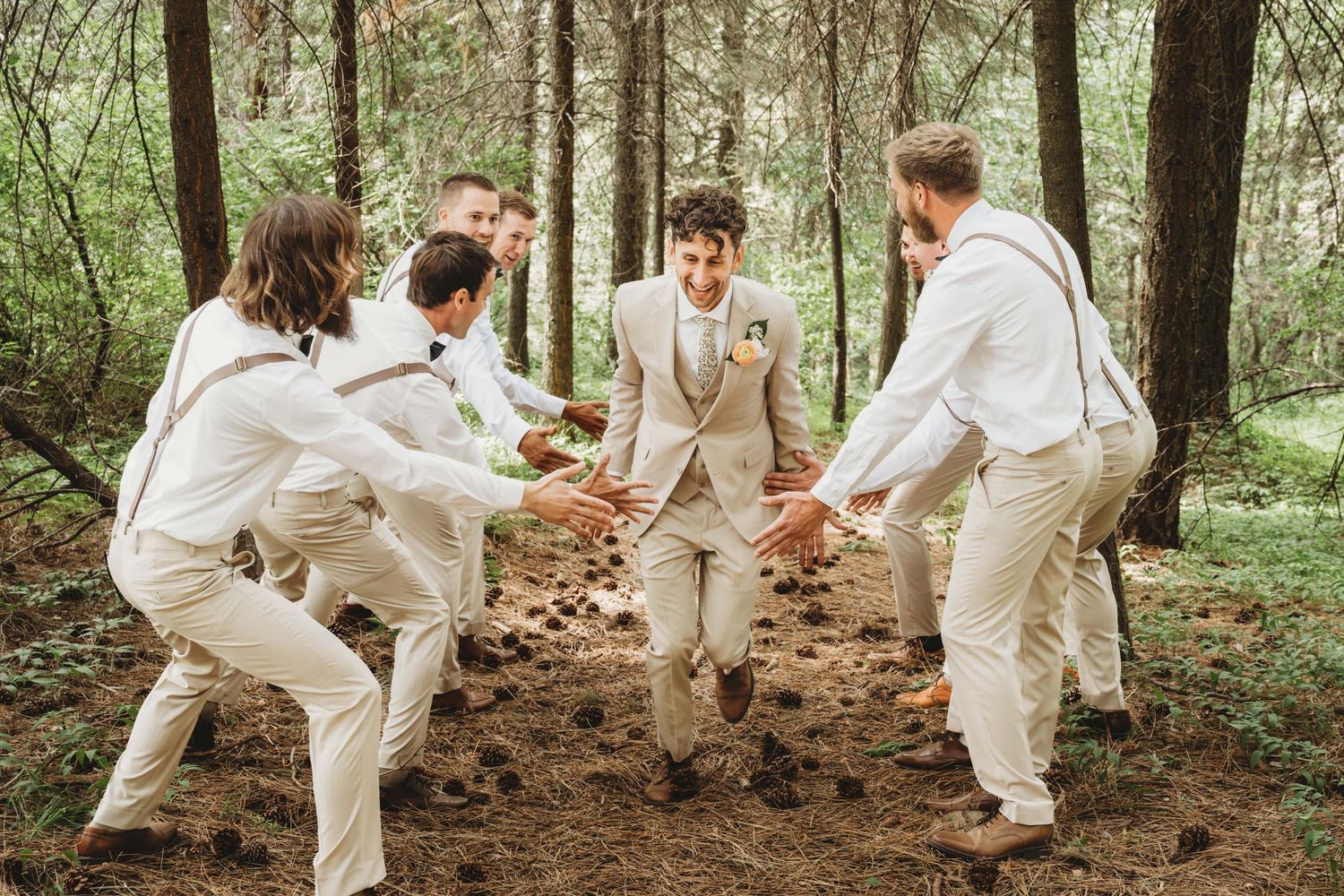 A groom running through a wooded area with his groomsmen clapping his hands as he runs by them.
