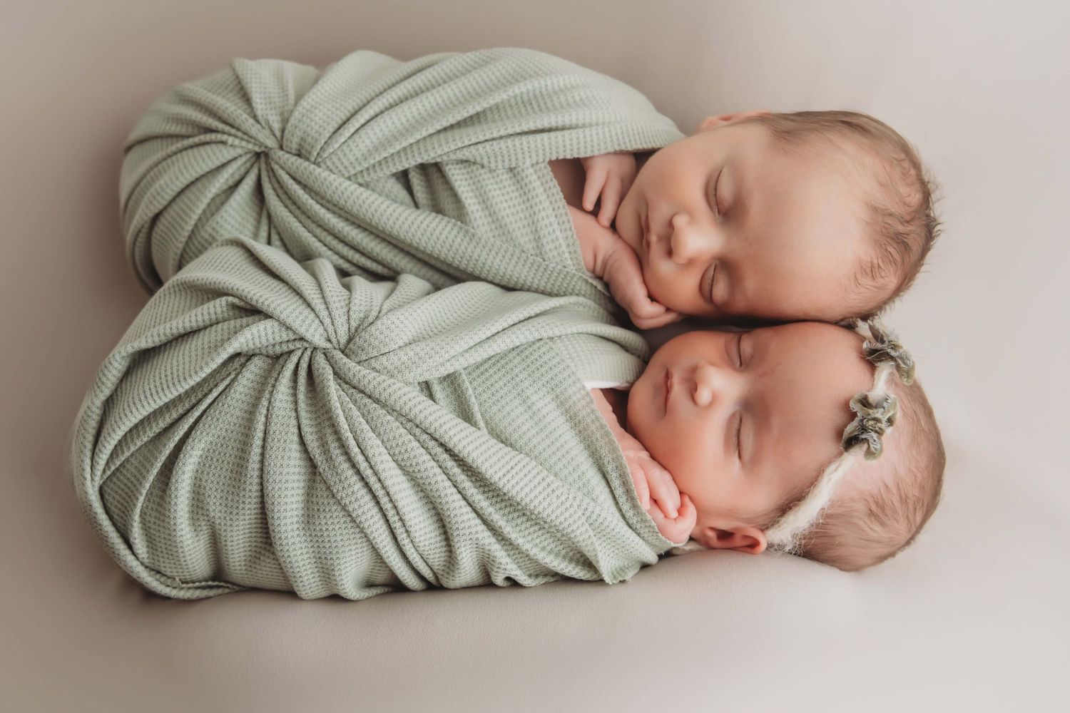 A newborn photo of twins sleeping together wrapped in sage green wraps.