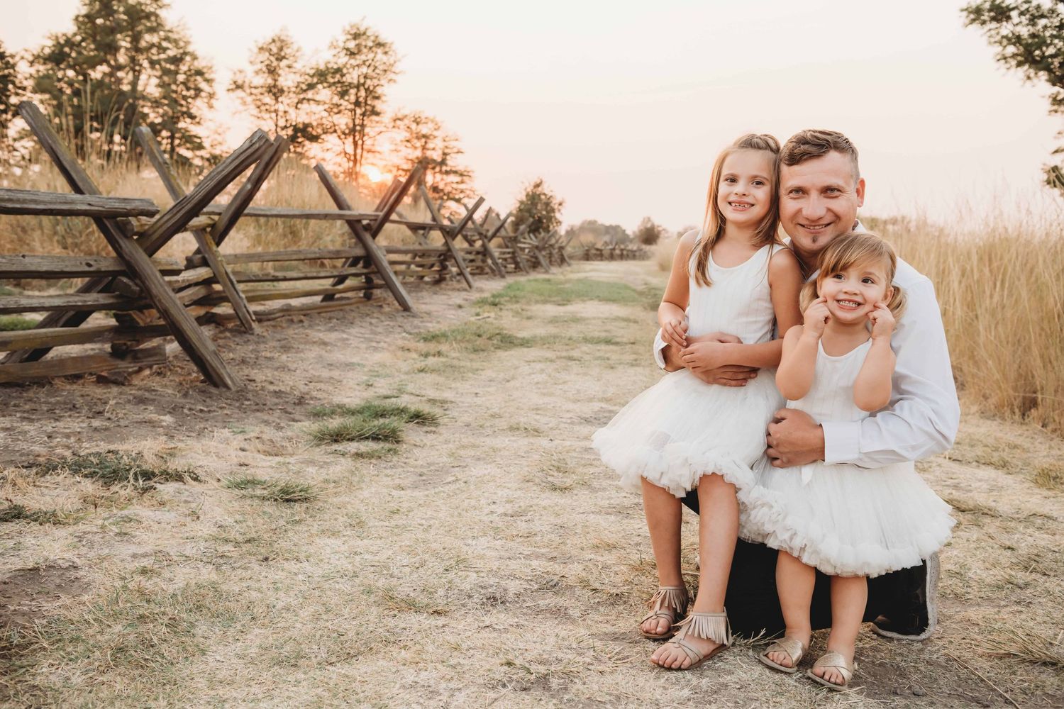 A father holding his two daughters with a fence and trees in the background.