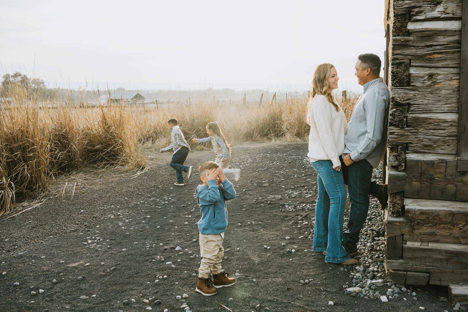 Children playing hide and seek while their mom and dad talk while leaning against a log building.