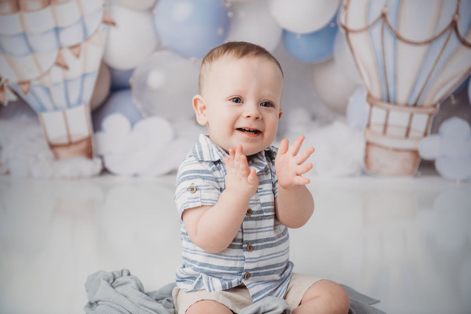 A baby sitting in front of a blue hot air balloon backdrop clapping his hands and smiling.