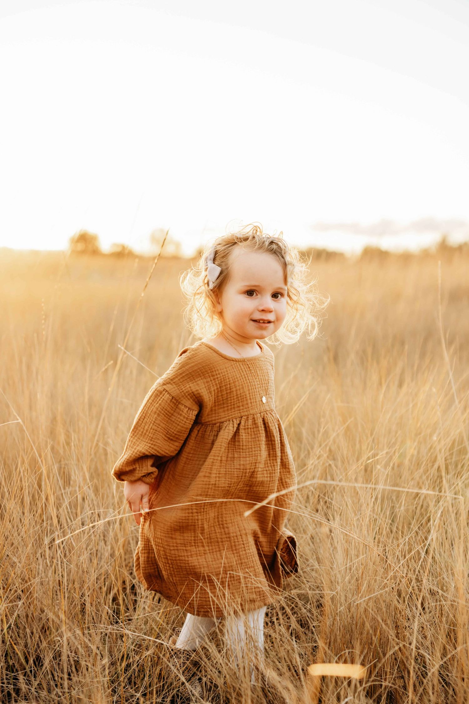 A young girl walking through a golden field with the sun shining through her curly hair.