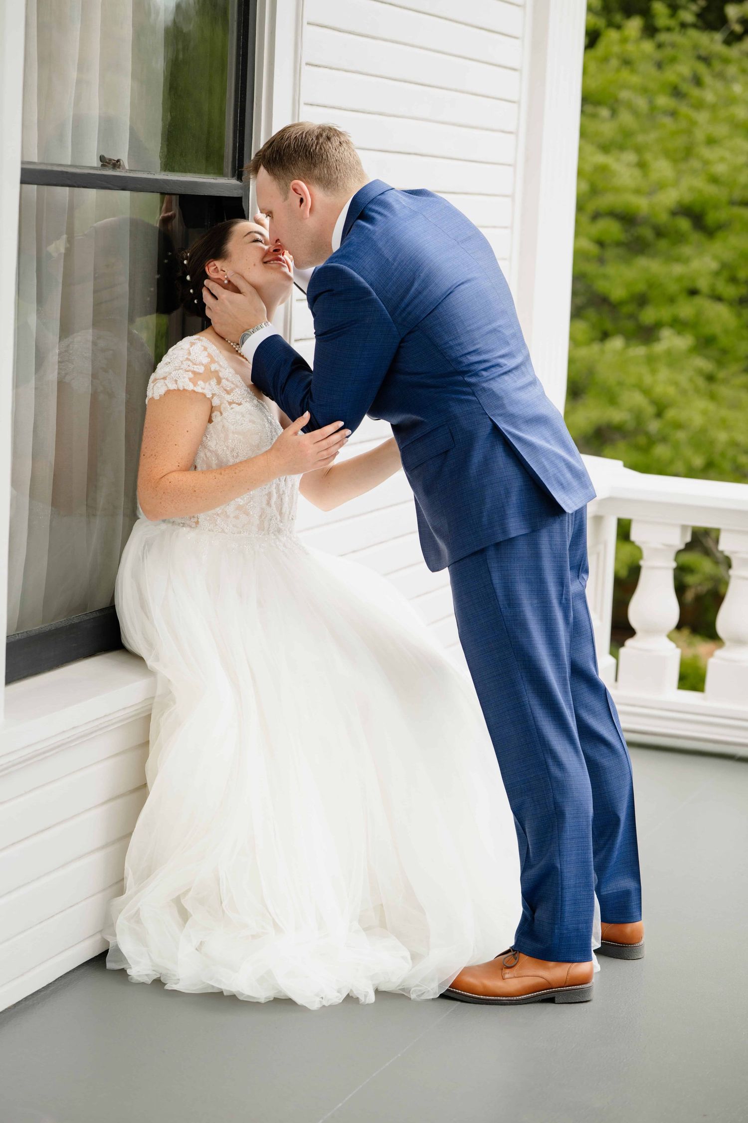 A woman sitting against a windowsill on a balcony while her groom gives her a kiss.