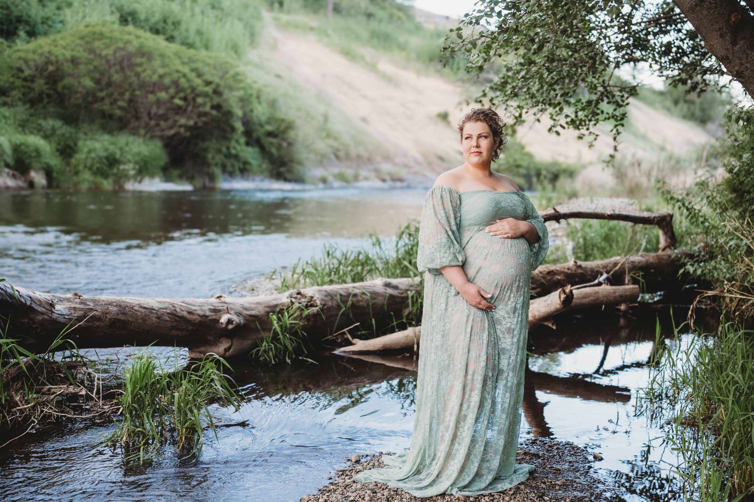 A pregnant woman standing in a shallow creek and looking off in the distance while holding her belly.