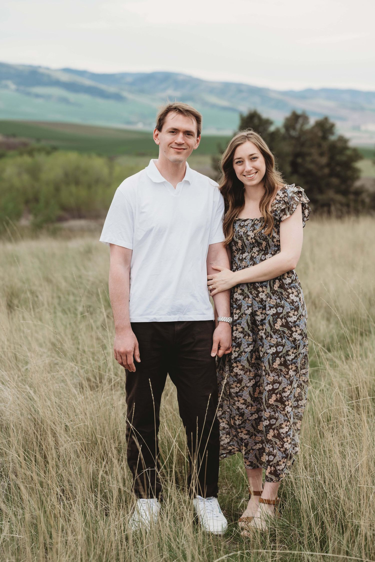 A couple standing in a dry grassy field holding hands and smiling at the camera.