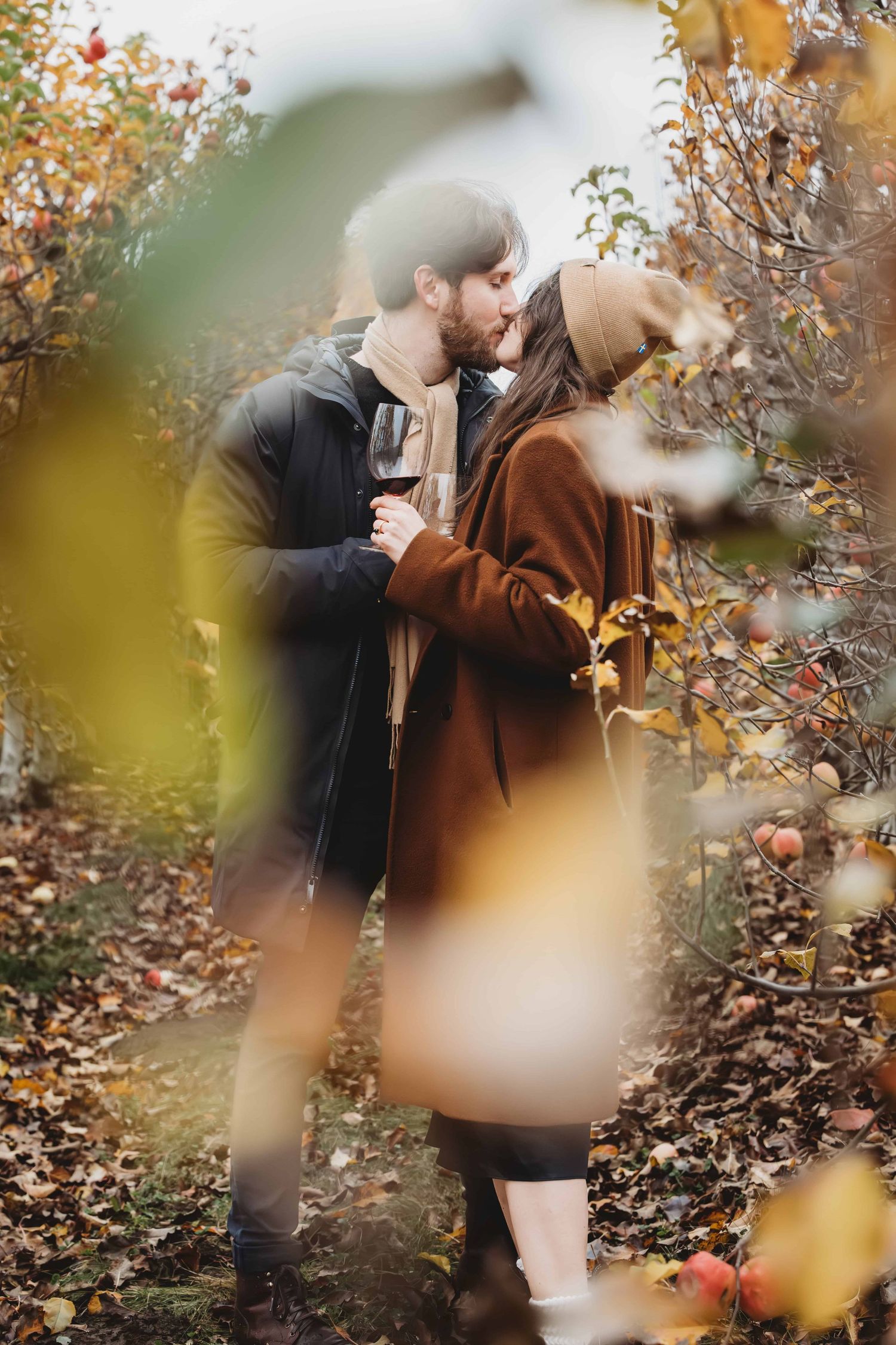 A couple photographed through some apple tree leaves, kissing and holding wine glasses just after getting engaged.
