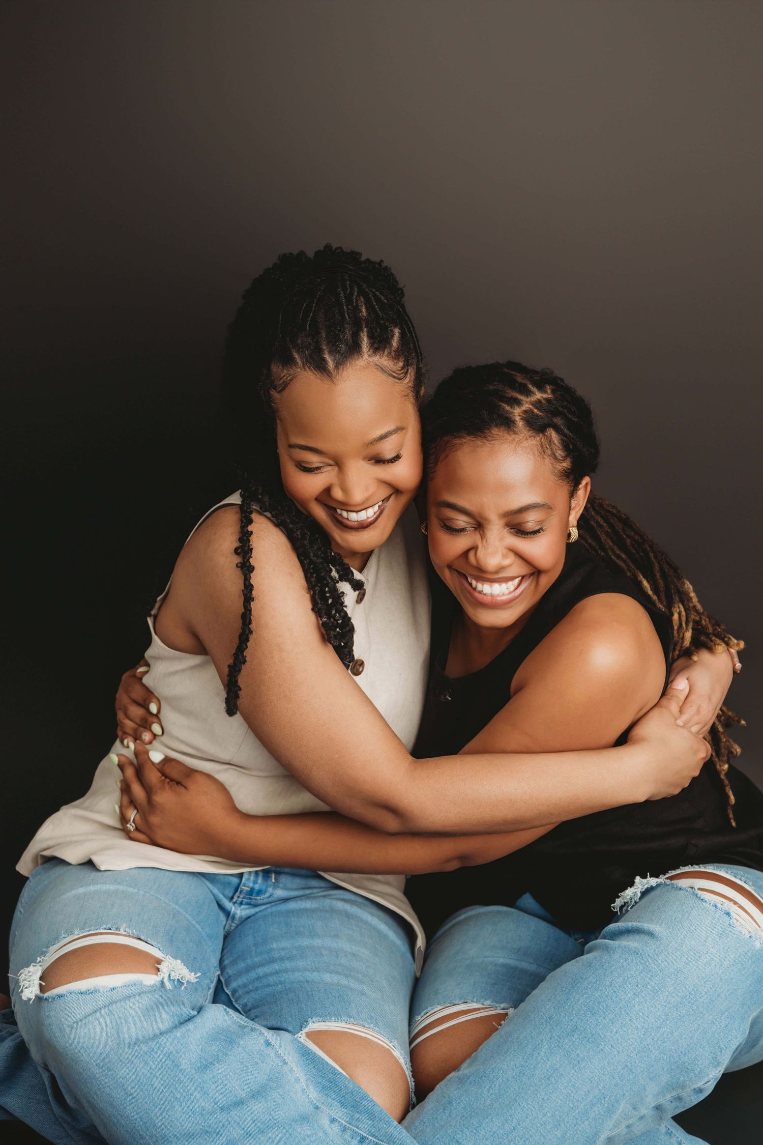 Two adult sisters sit on a black backdrop hugging each other for a candid studio portrait in Walla Walla.