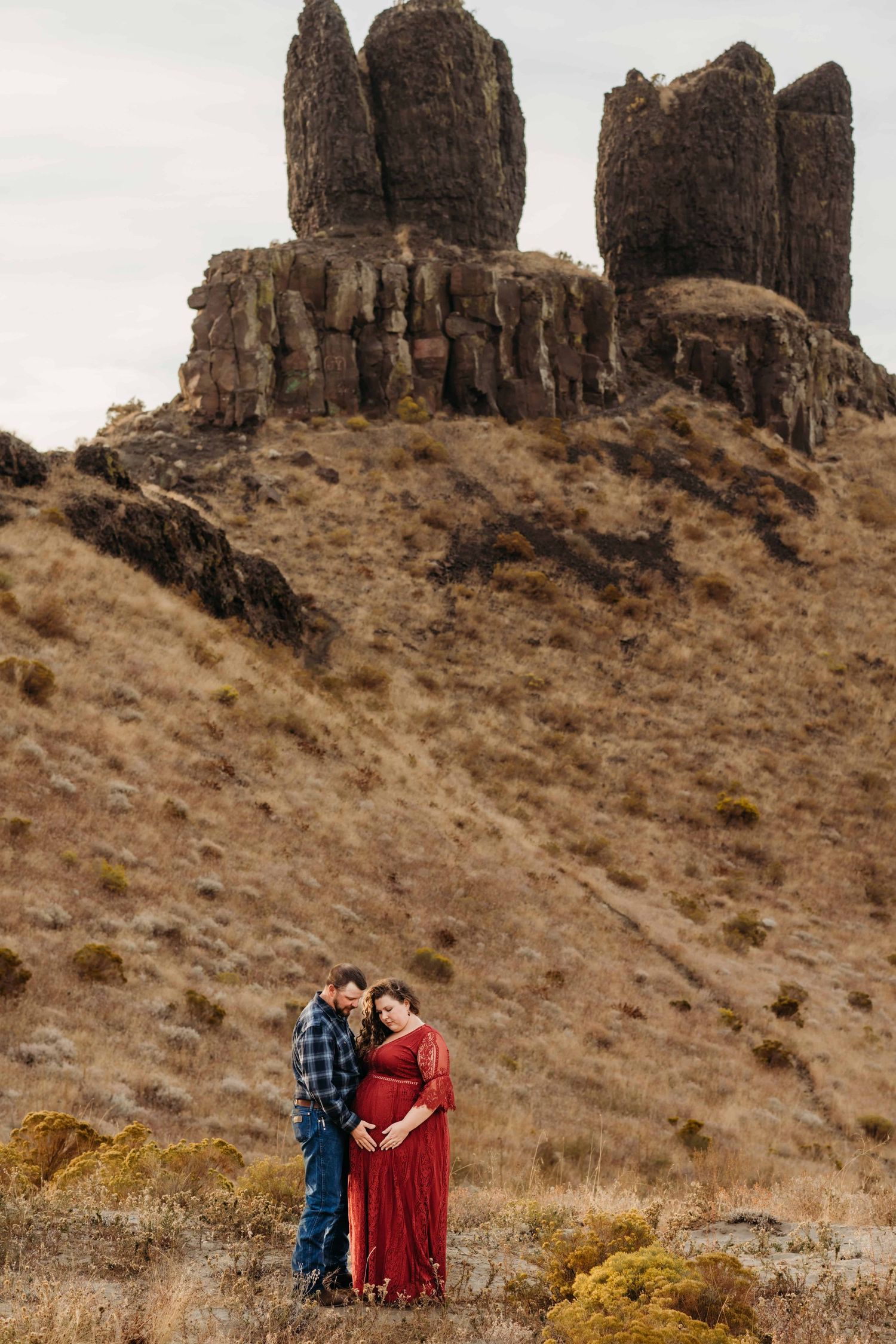 A portrait of a couple looking down at the baby bump with the Twin Sisters Rock formation in the background.