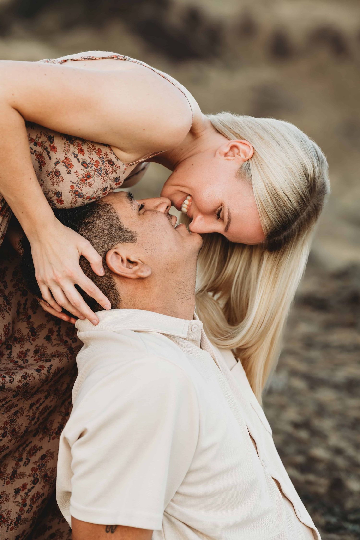 A woman standing above a man sitting down on a rock giving him a kiss.