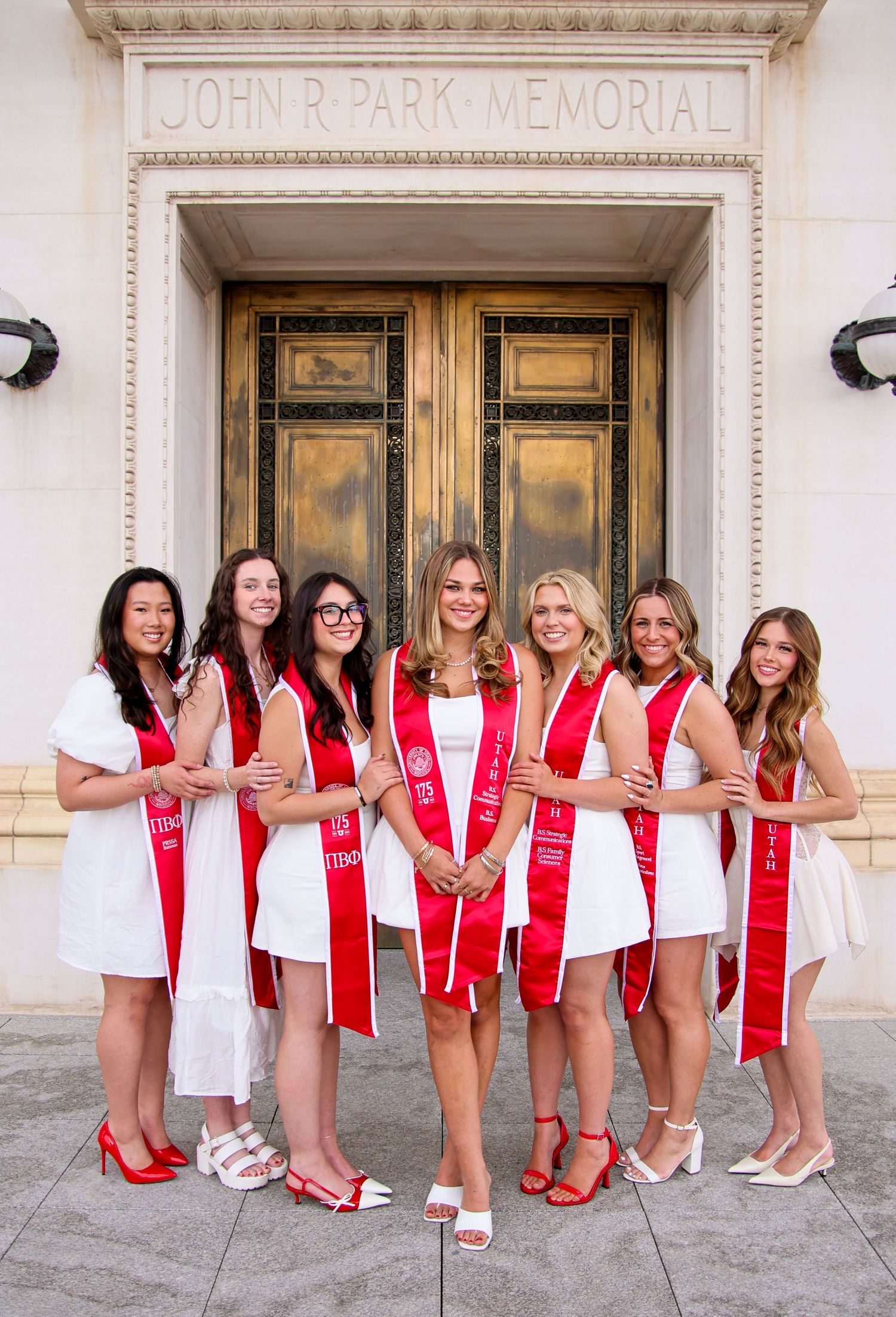 Graduates in white dresses and red sashes pose together outside John R Park Memorial building.