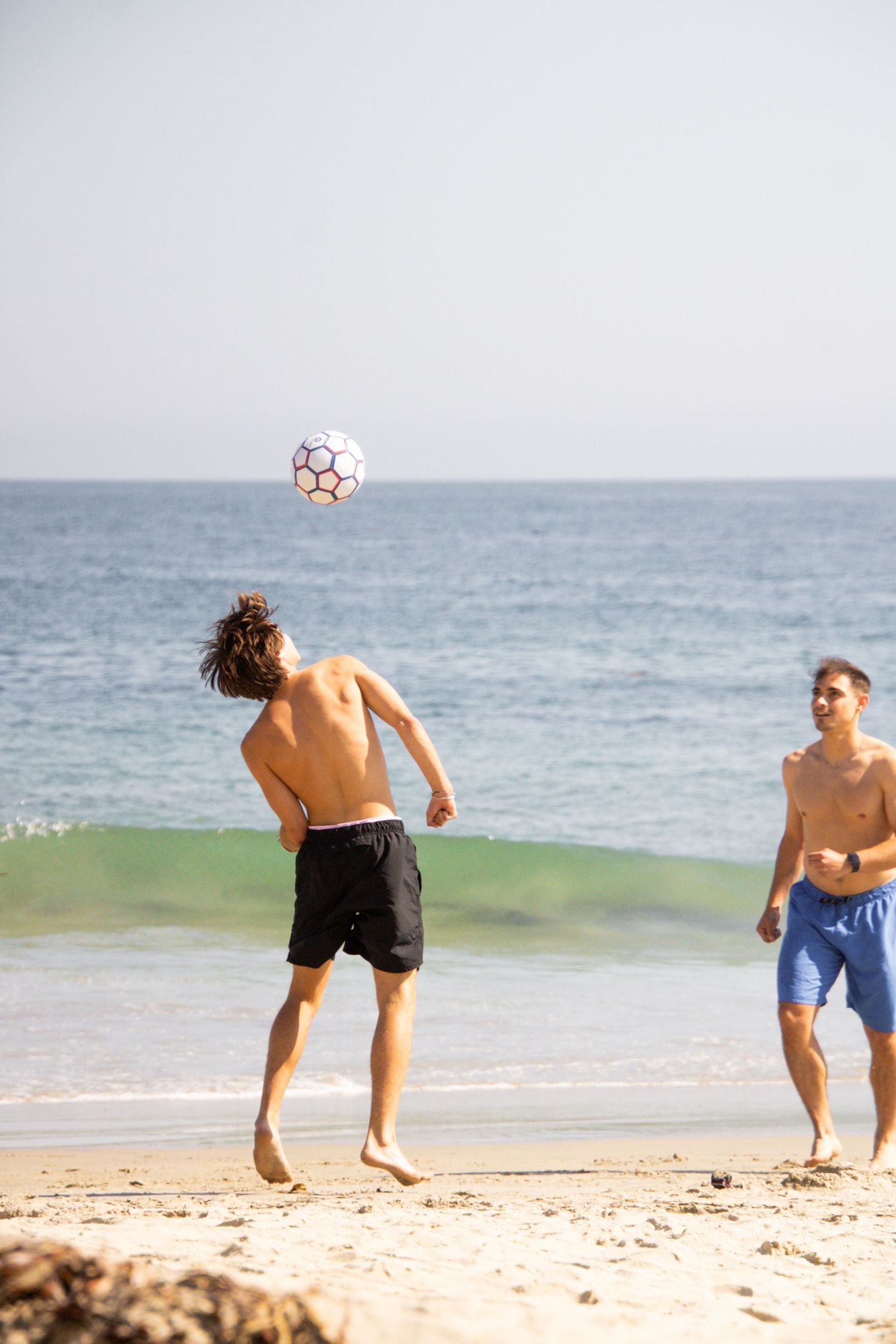 Friends play soccer together on a sandy beach with waves crashing behind them.