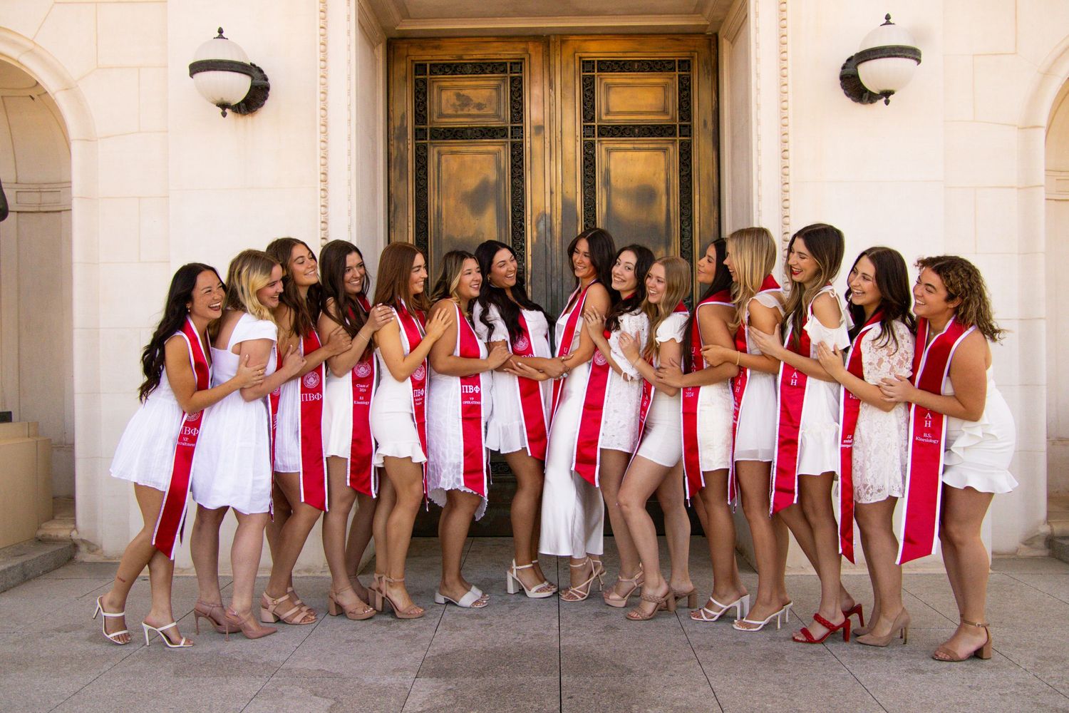 Large group of graduates in red stoles and black gowns pose together outside building.