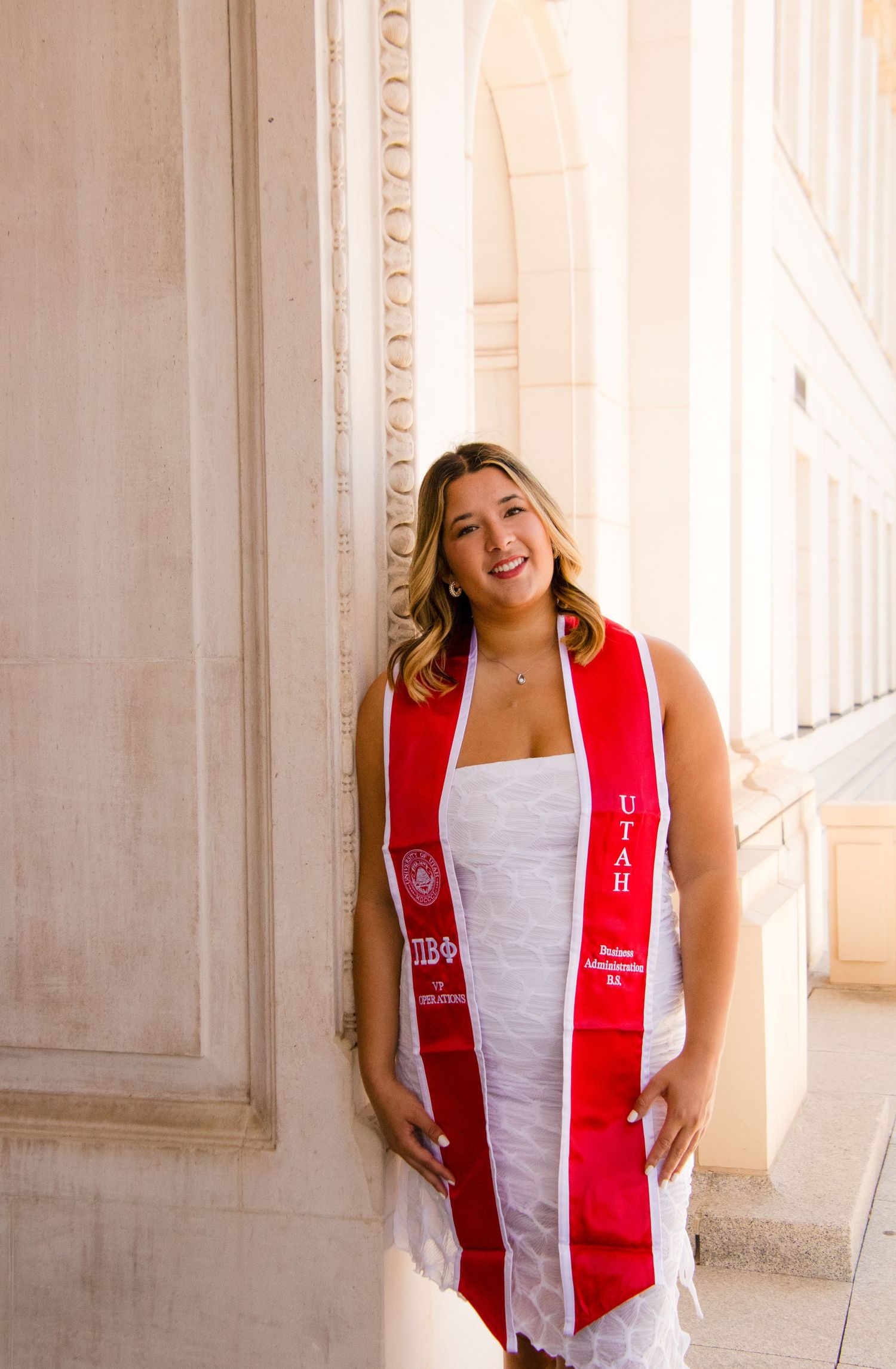Graduate in white dress and red sorority stole celebrates joyfully against stone pillars.