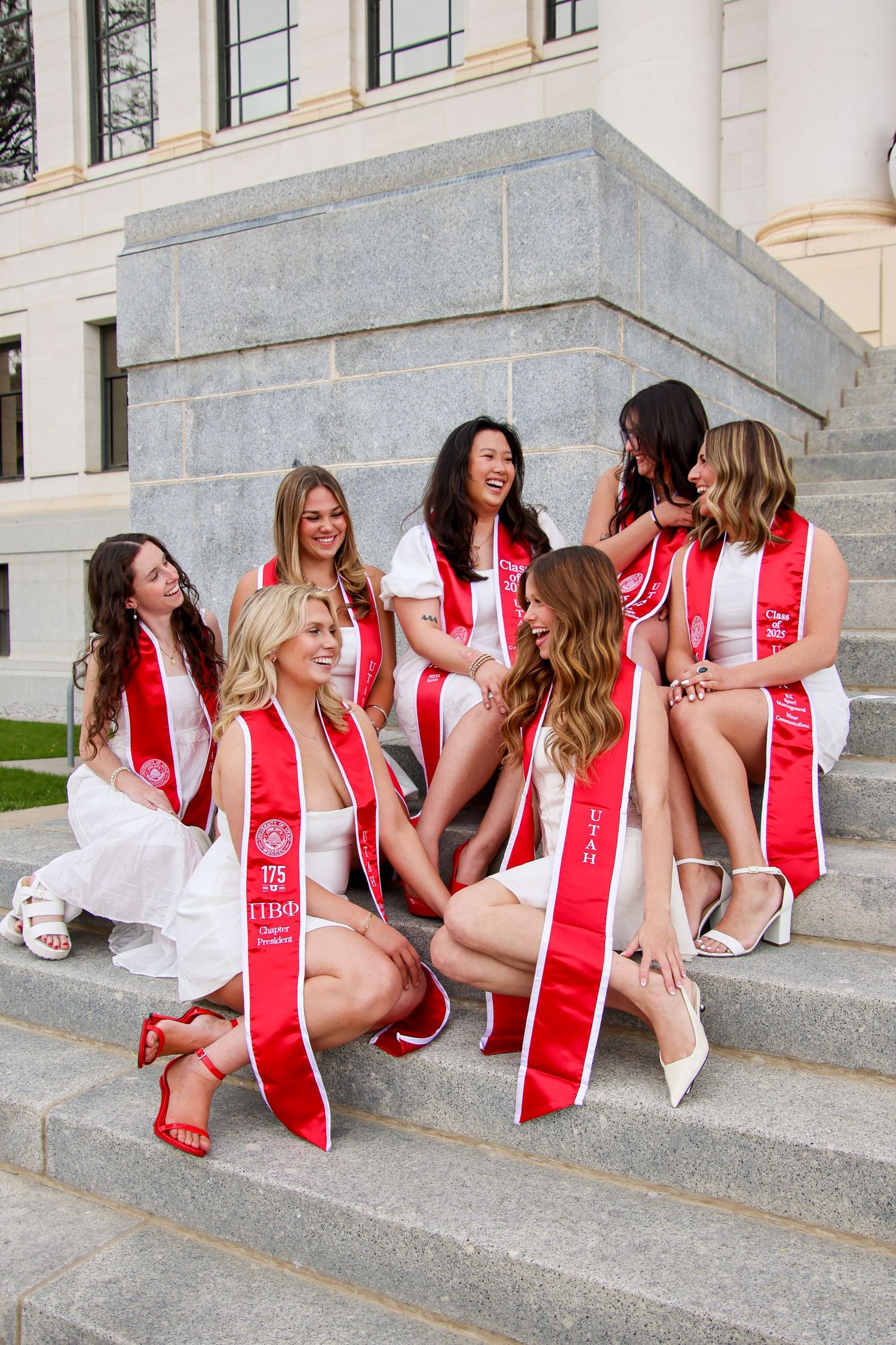 Group of graduates in white dresses and red stoles sitting on stone steps during graduation photo session.