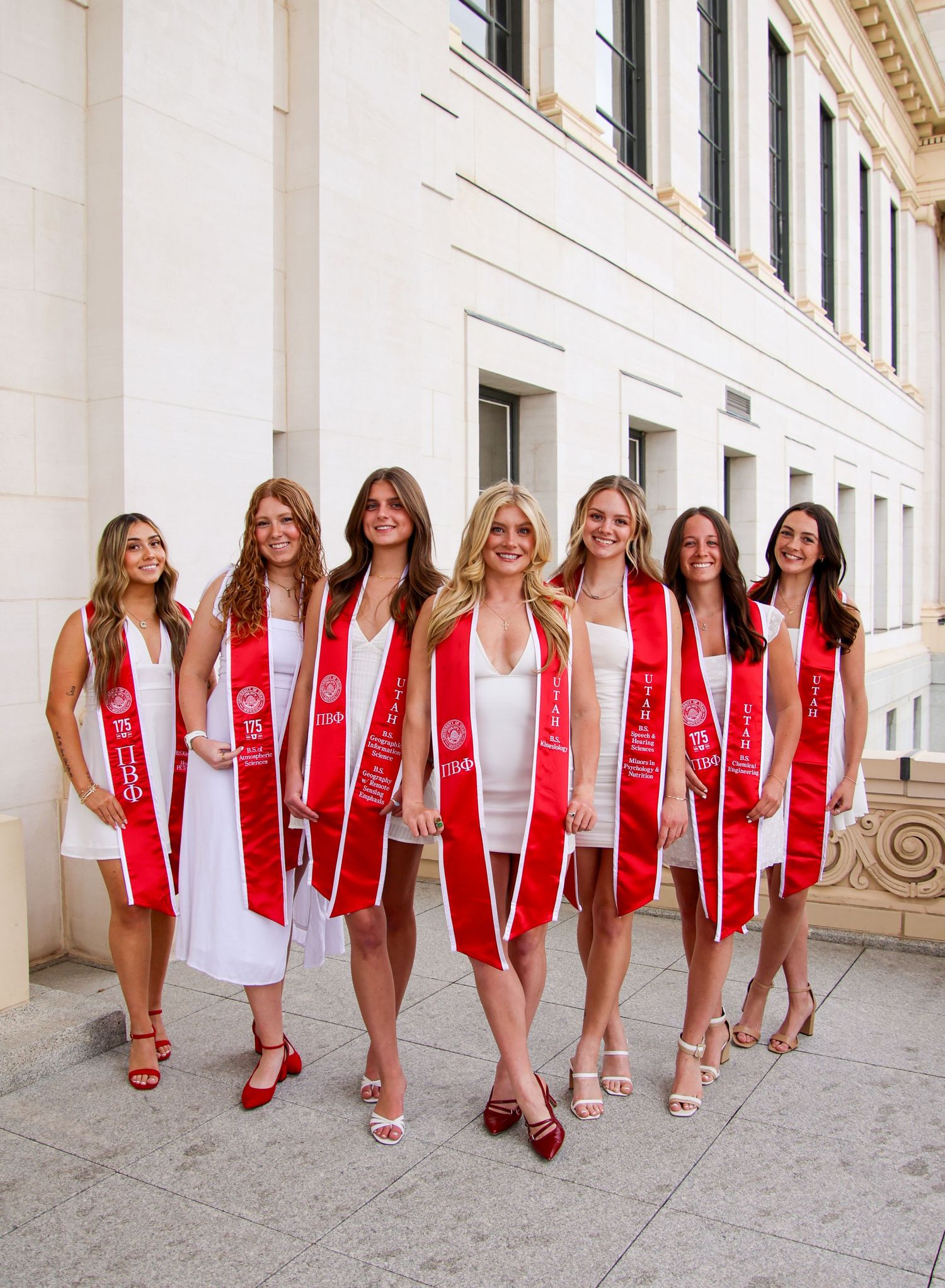 Group photo of graduates in white dresses and red sashes posing together on steps of a building.