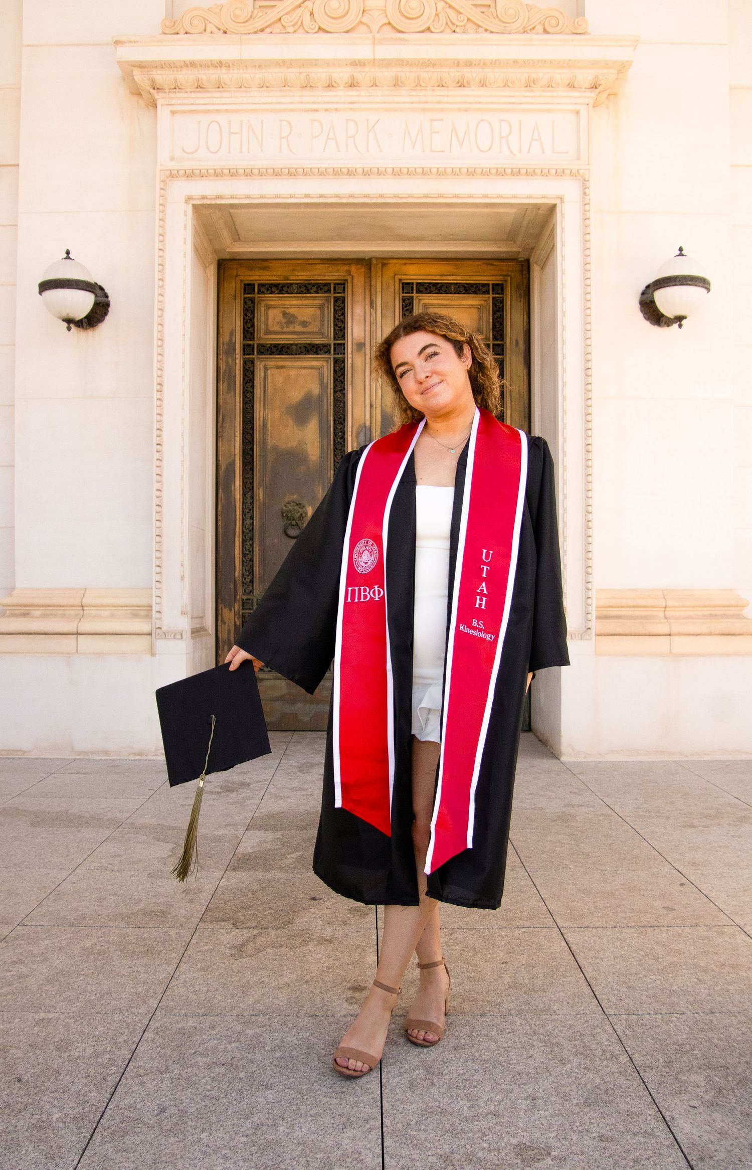 Graduate in black gown and red stole poses playfully outside ornate building entrance.
