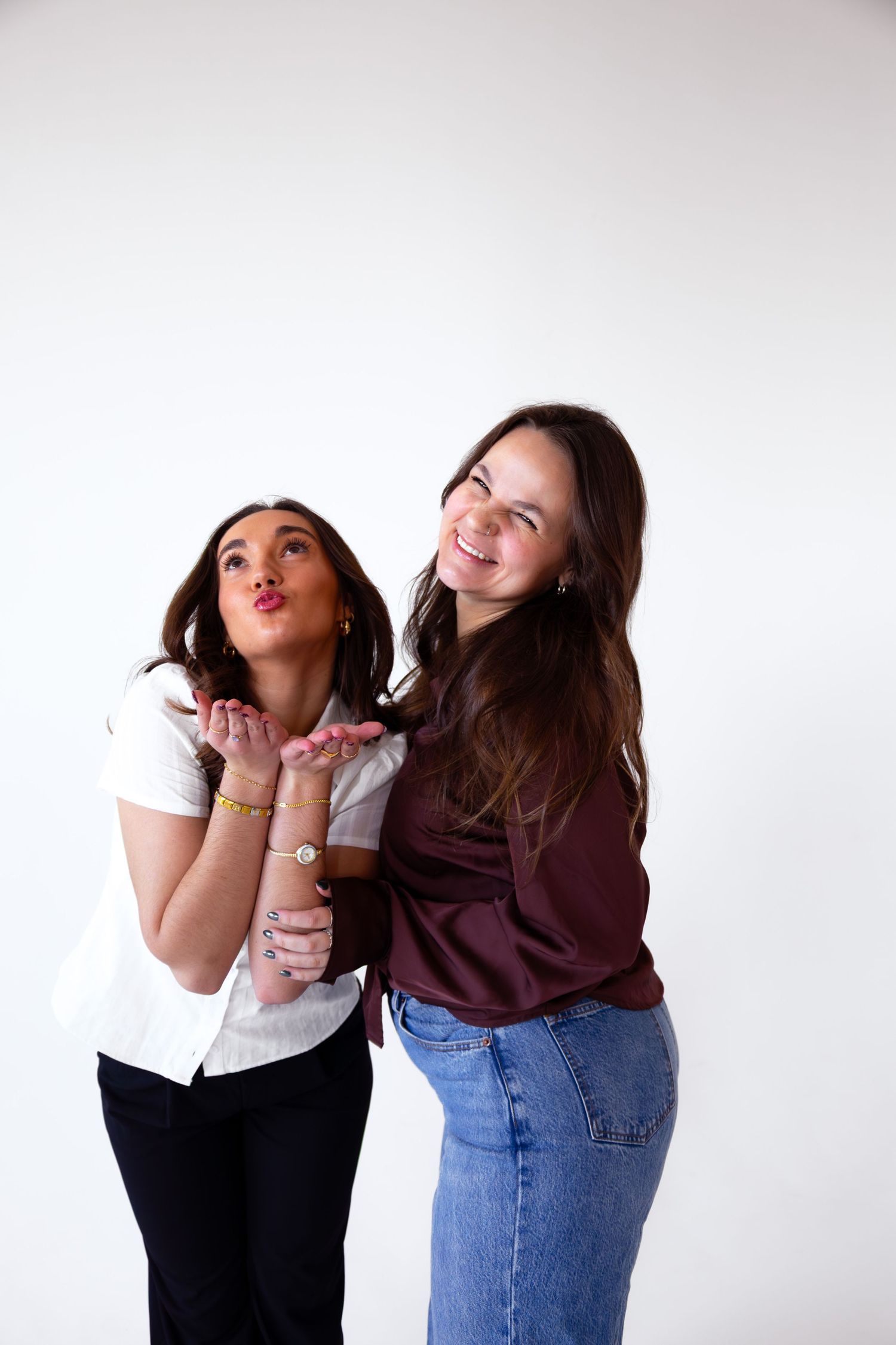 Two friends pose playfully together against a white background in casual outfits.