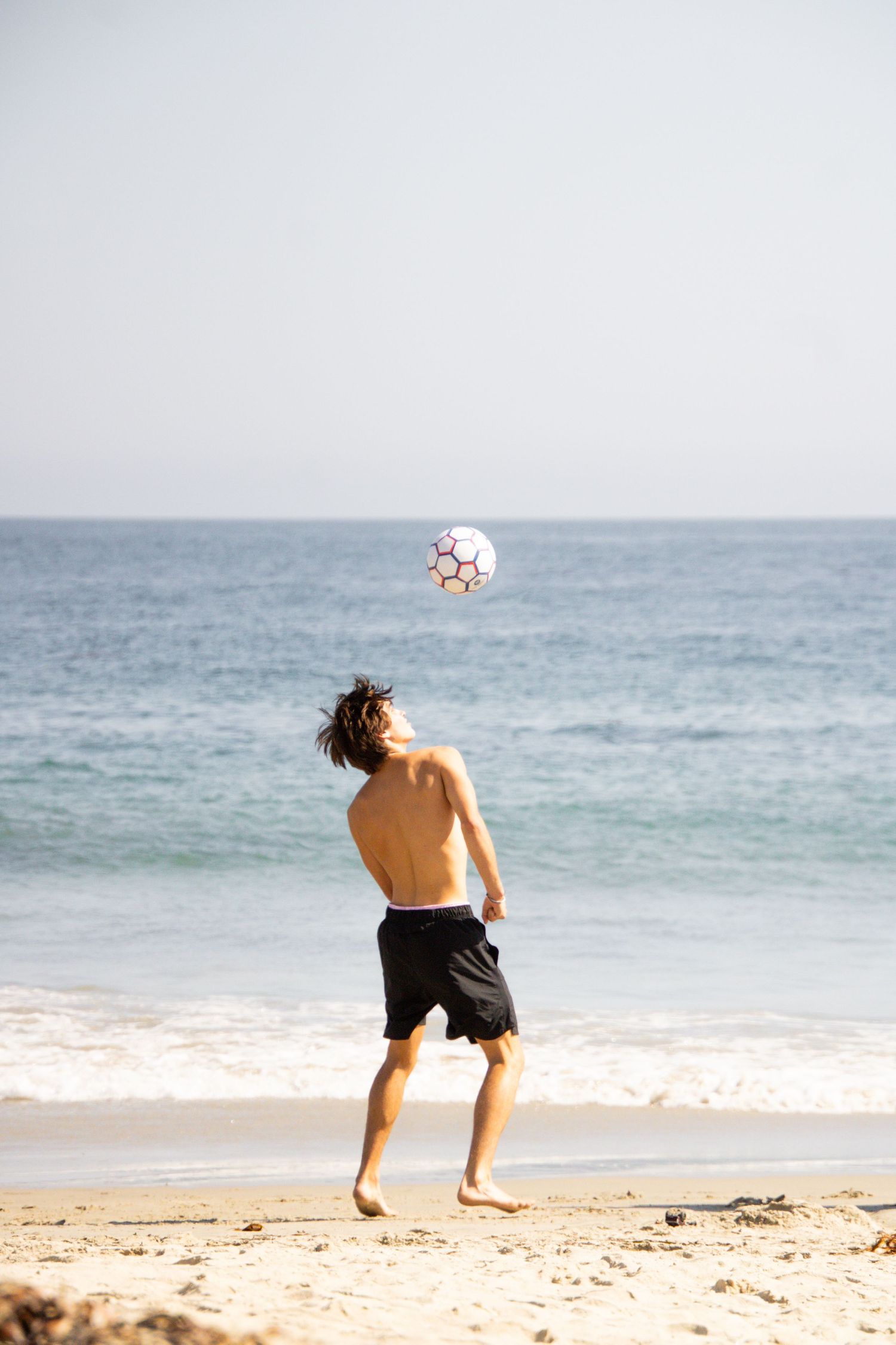 A beachgoer plays with a soccer ball on sandy shores beside the ocean.