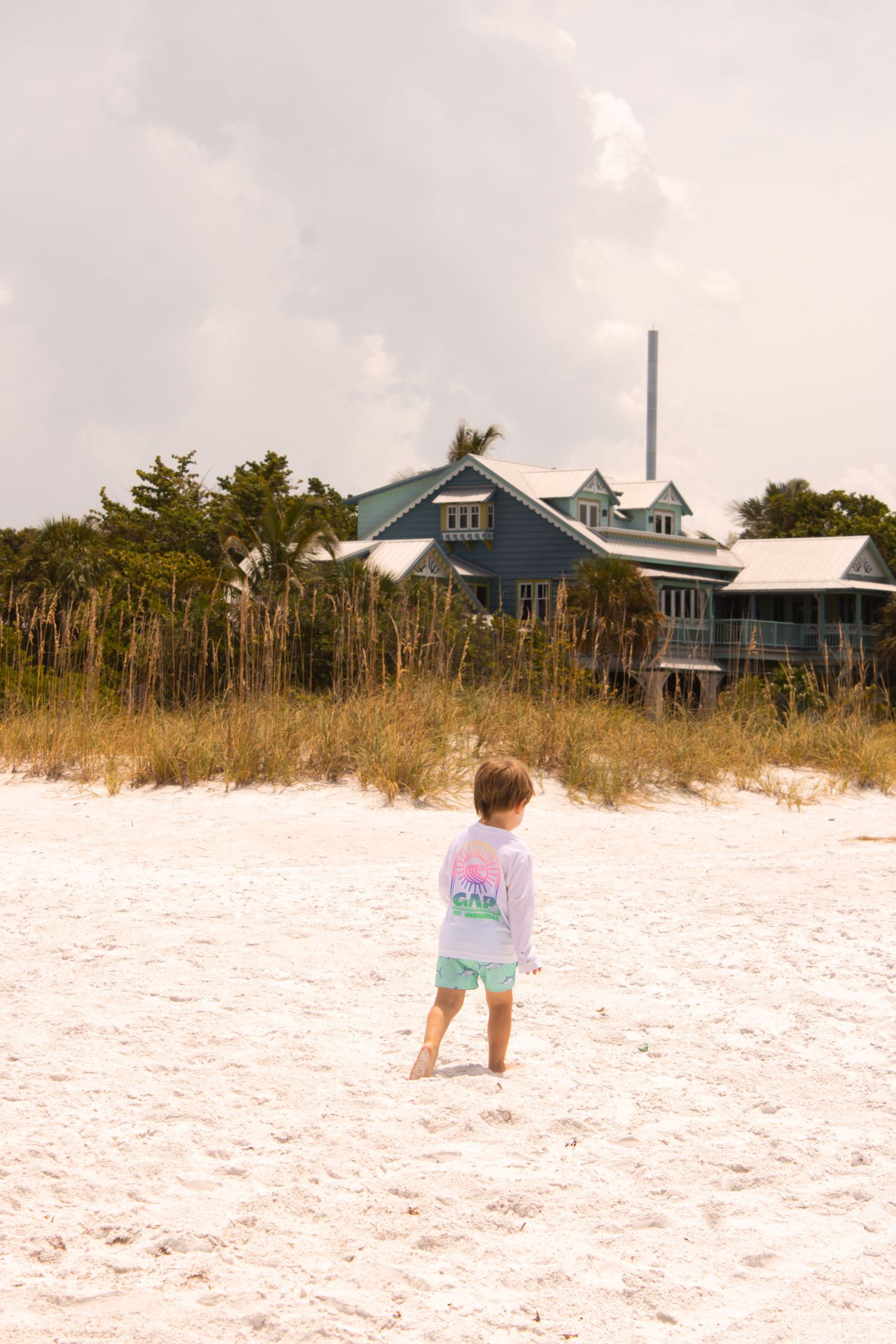 A small child plays in white sand with beach houses visible in the background.
