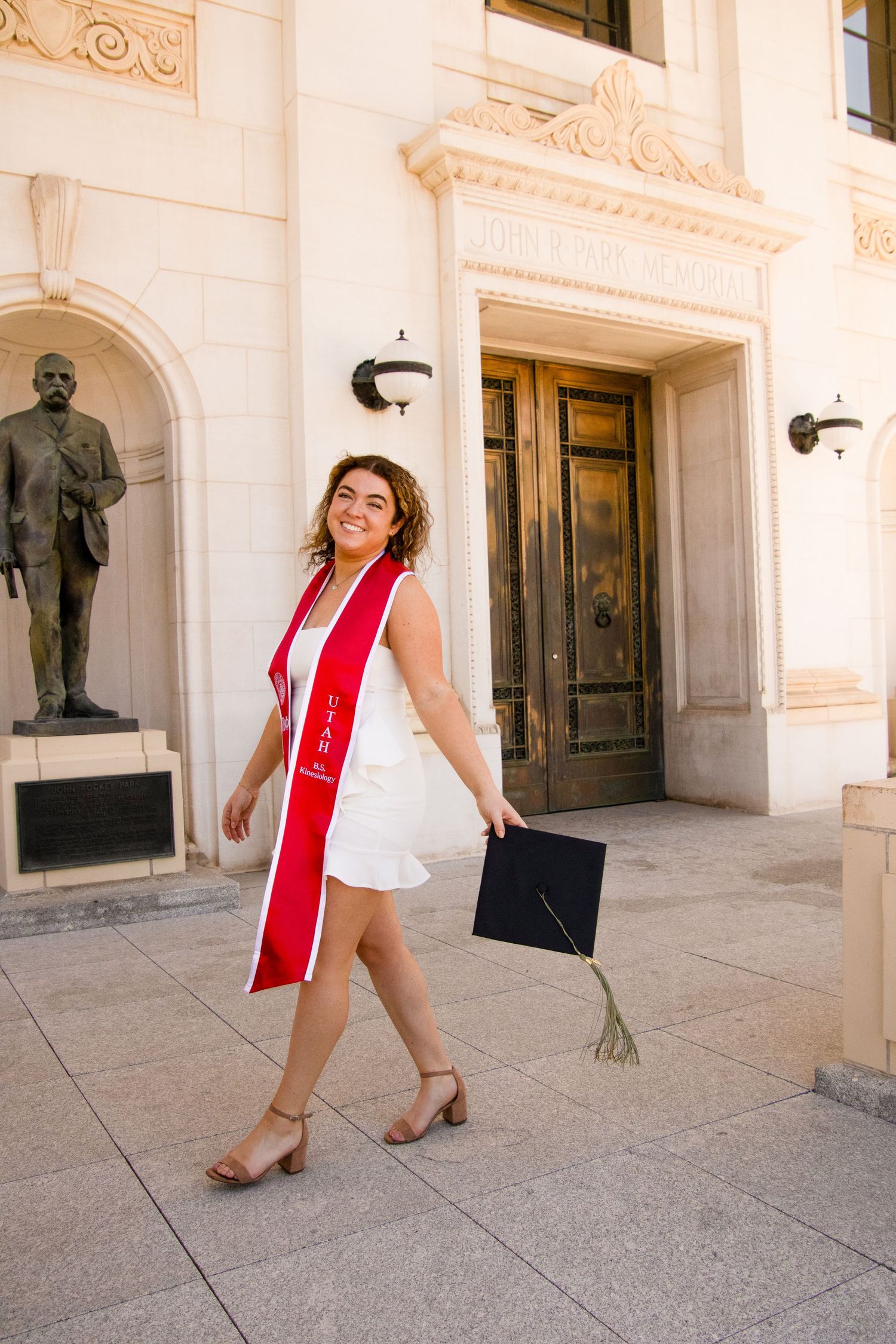 Graduate in white dress and red stole dances playfully on campus grounds.