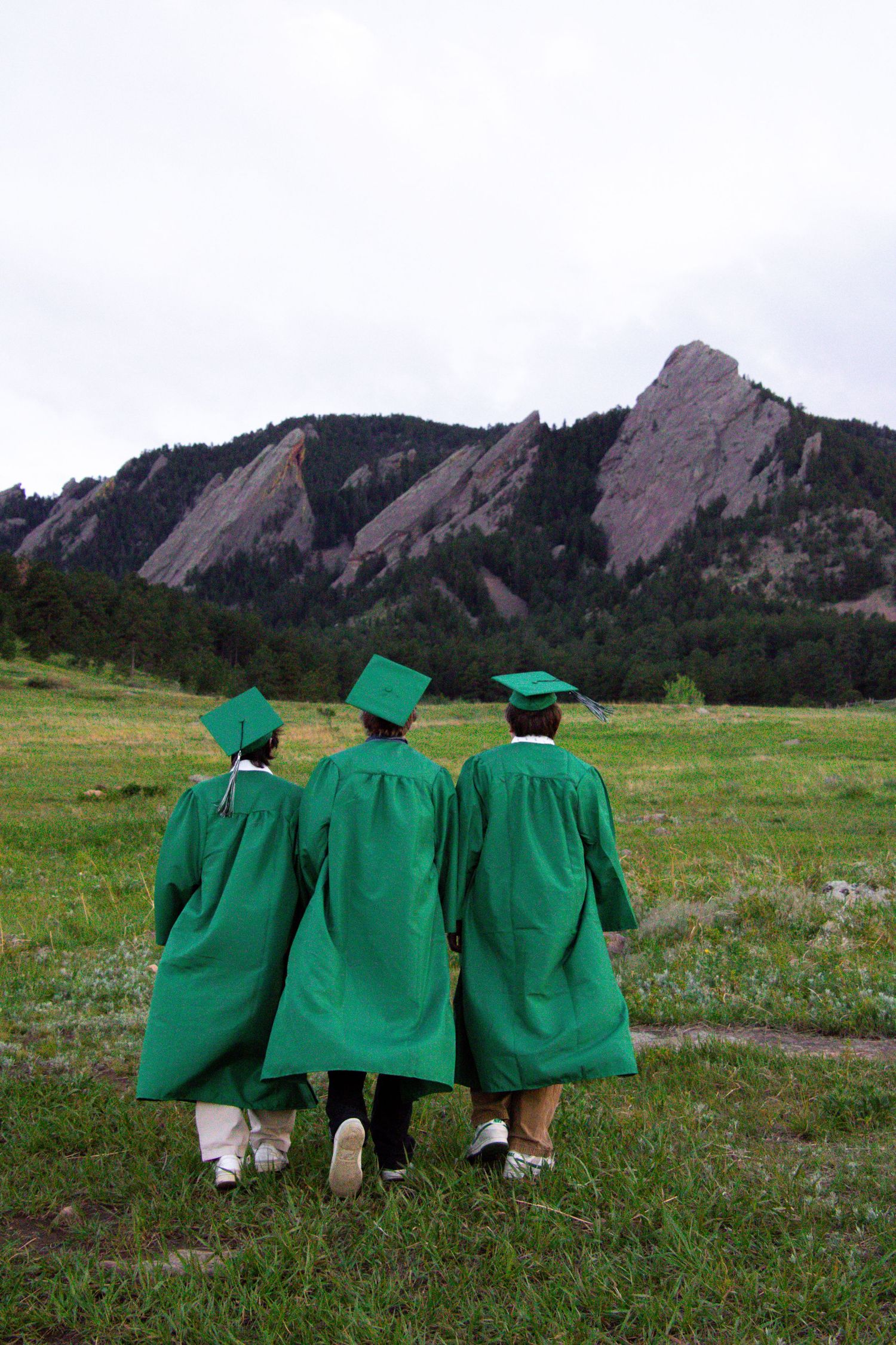 Three graduates in green robes pose with their backs turned against the backdrop of the Flatirons mountains in Boulder.