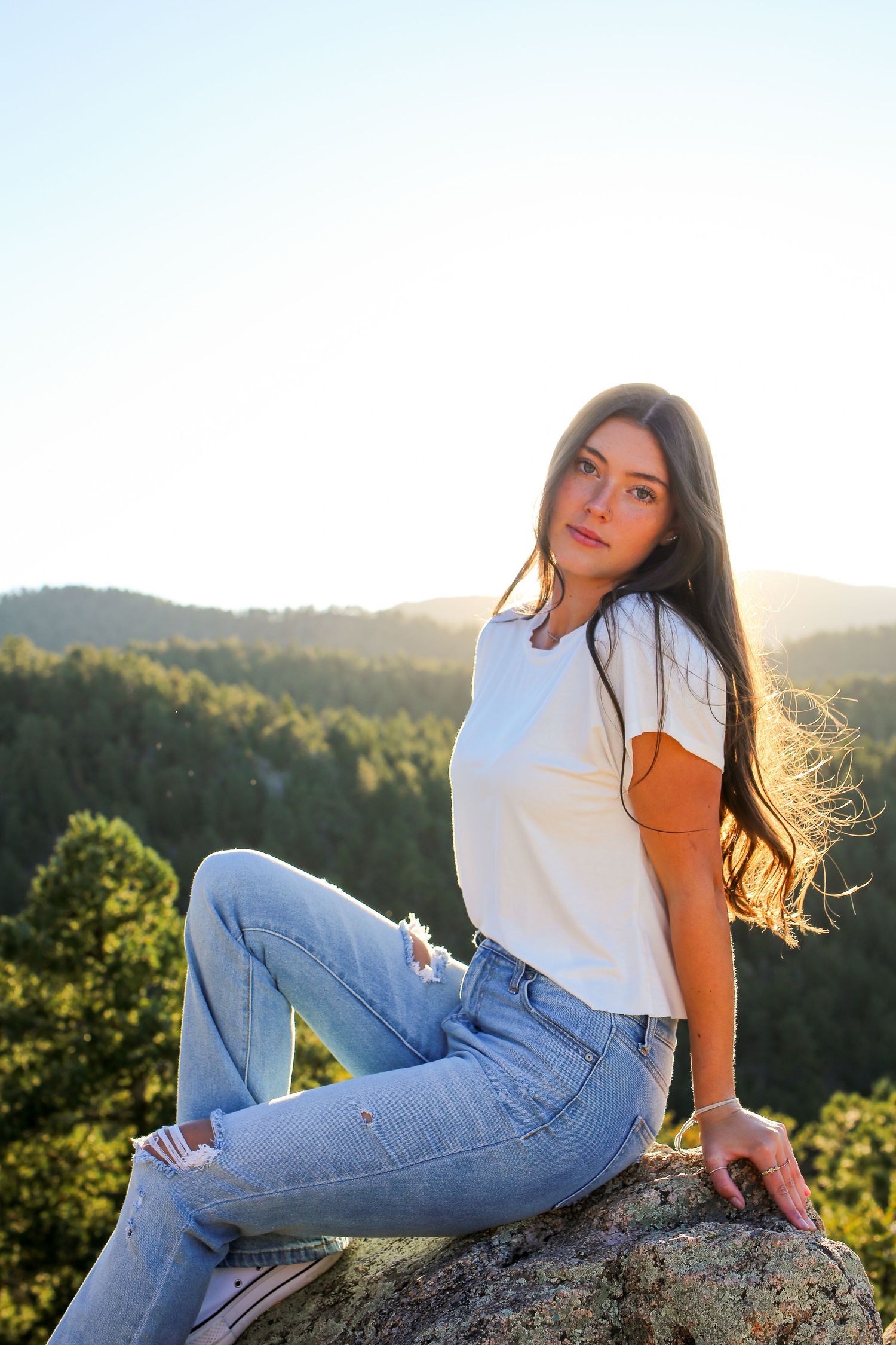 Portrait of someone in casual denim and white t-shirt sitting on rock with mountain vista behind.