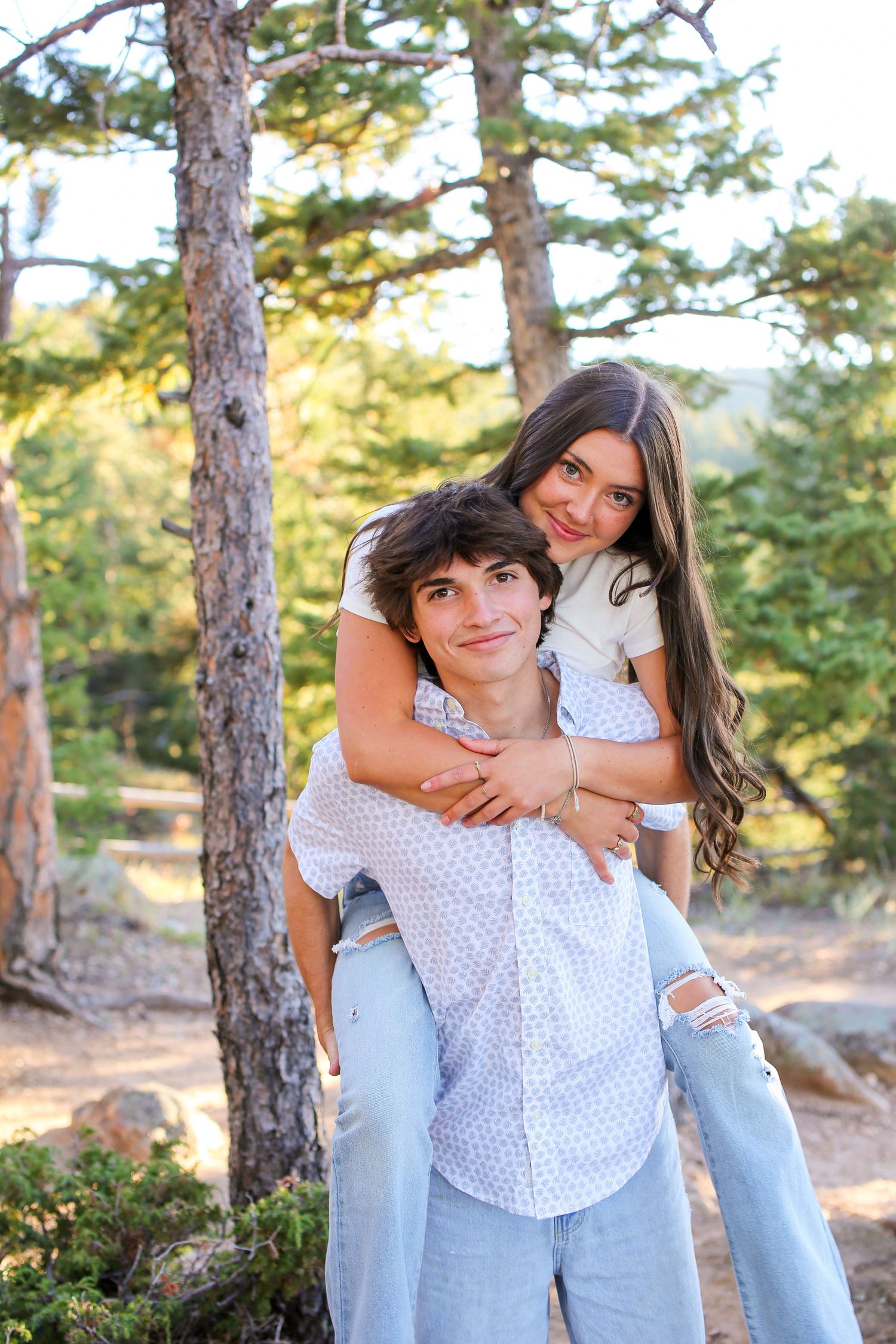 Playful couple in white and blue outfits posing among pine trees.