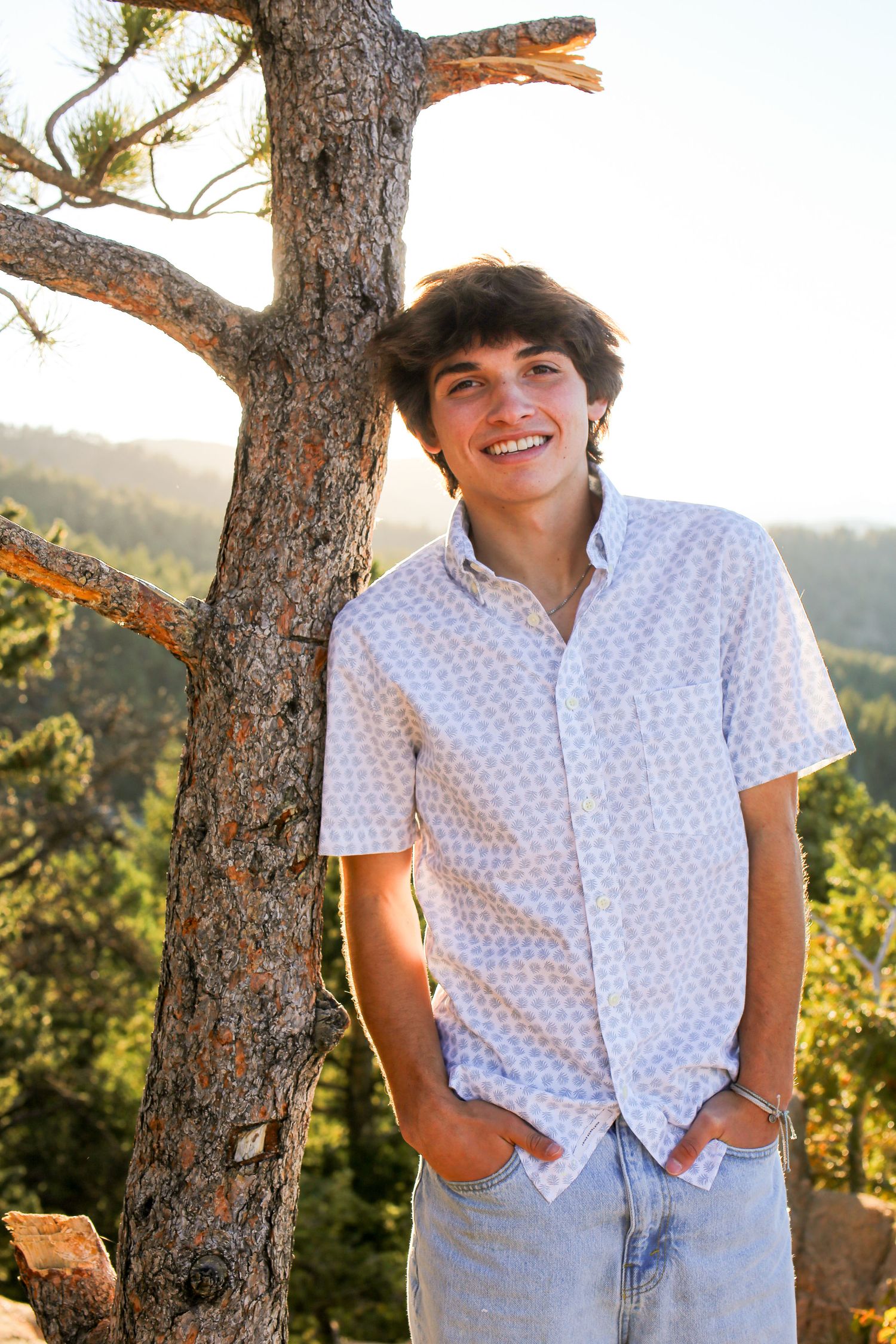 Summer portrait with someone in a white button-down shirt posing next to a pine tree with mountains in background.