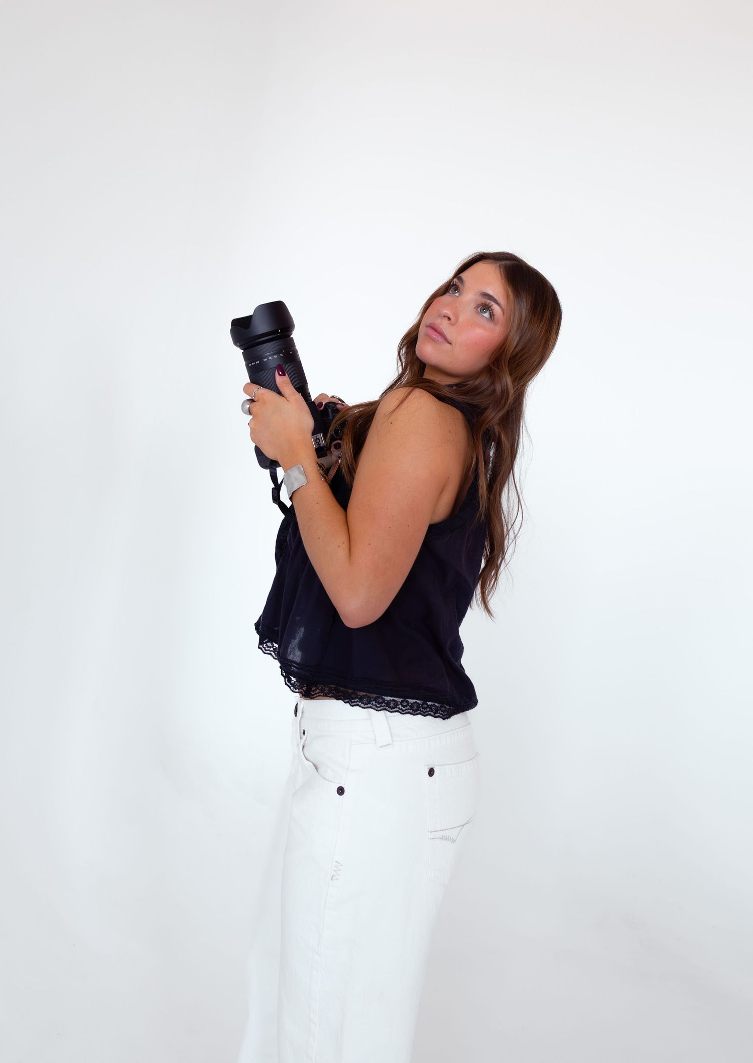 A photographer in black and white attire poses playfully with a camera against a white backdrop.