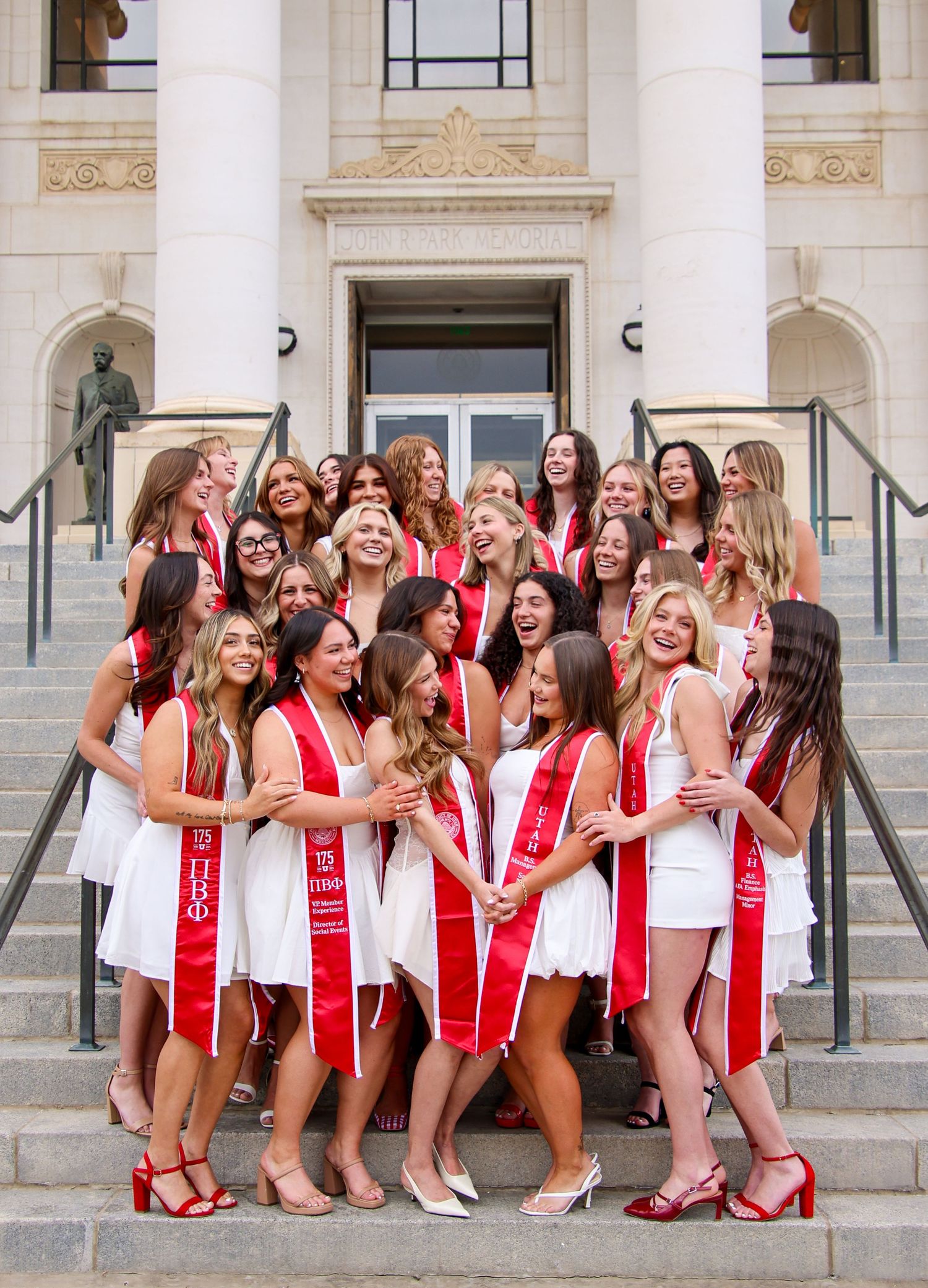 Celebratory group photo with red sashes and white dresses in front of a grand building entrance.