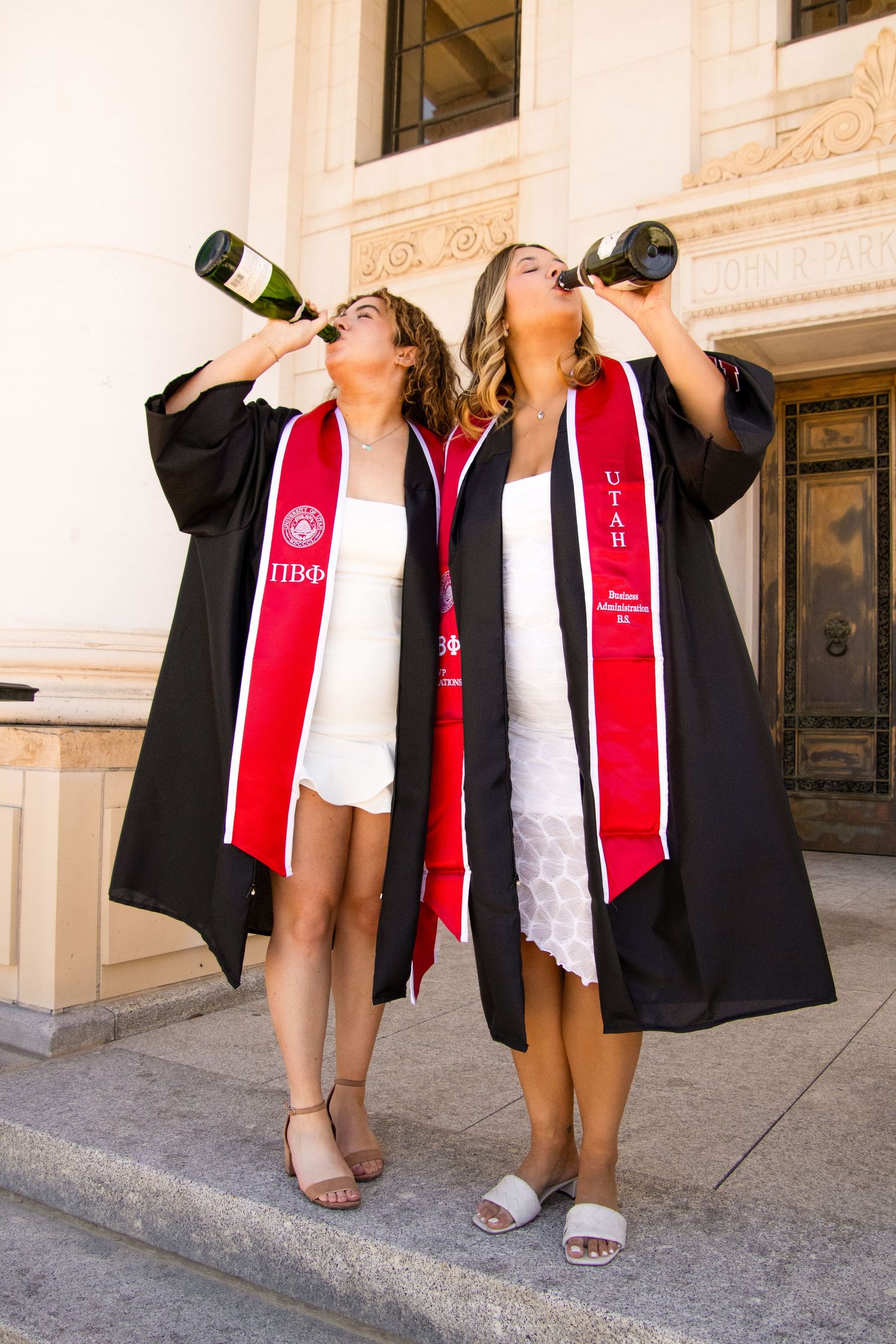 Graduates in graduation attire celebrate by popping champagne bottles on campus grounds.