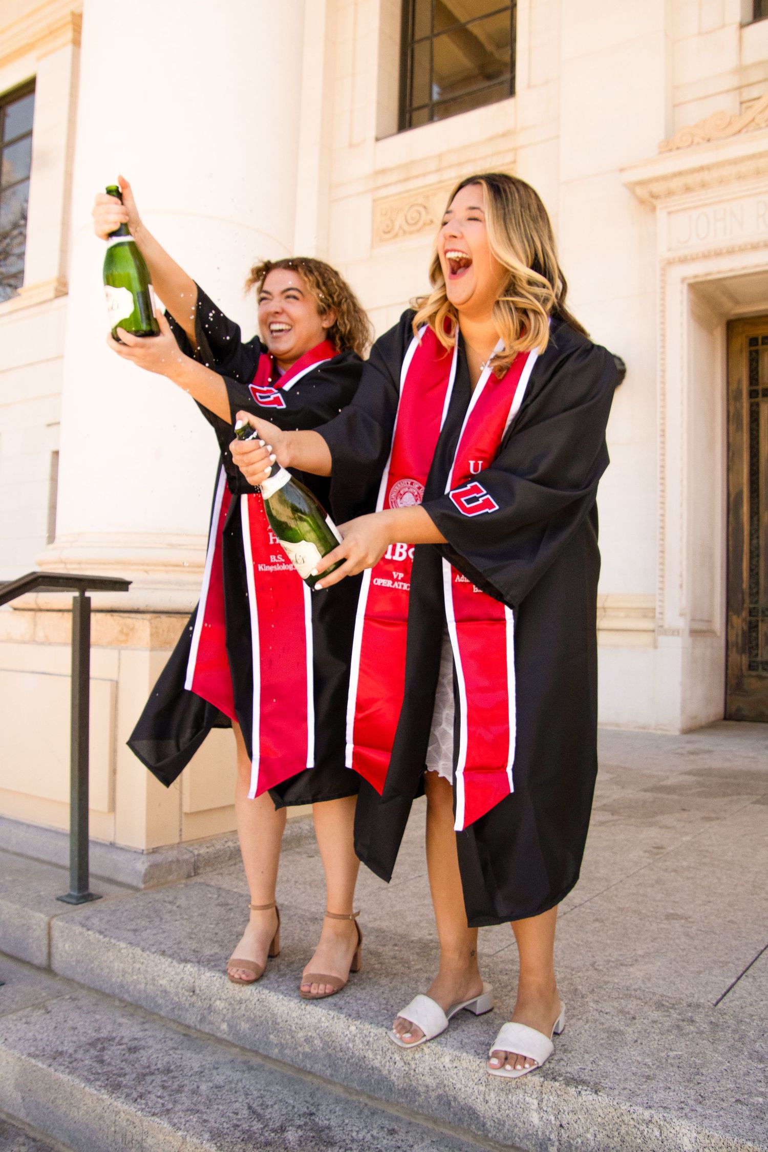 Graduates in black gowns and red stoles celebrate with champagne outside building.